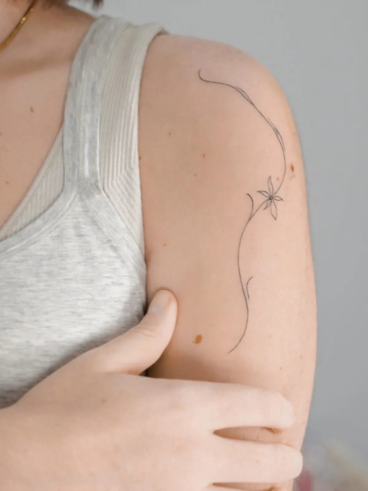 Brazo de una persona con un delicado tatuaje de enredadera, una fina línea con una pequeña flor, en el hombro.