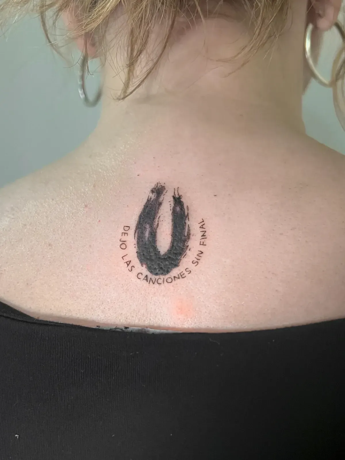Tatuaje de una herradura negra con texto en español, en la parte superior de la espalda de una persona, cerca del cuello.