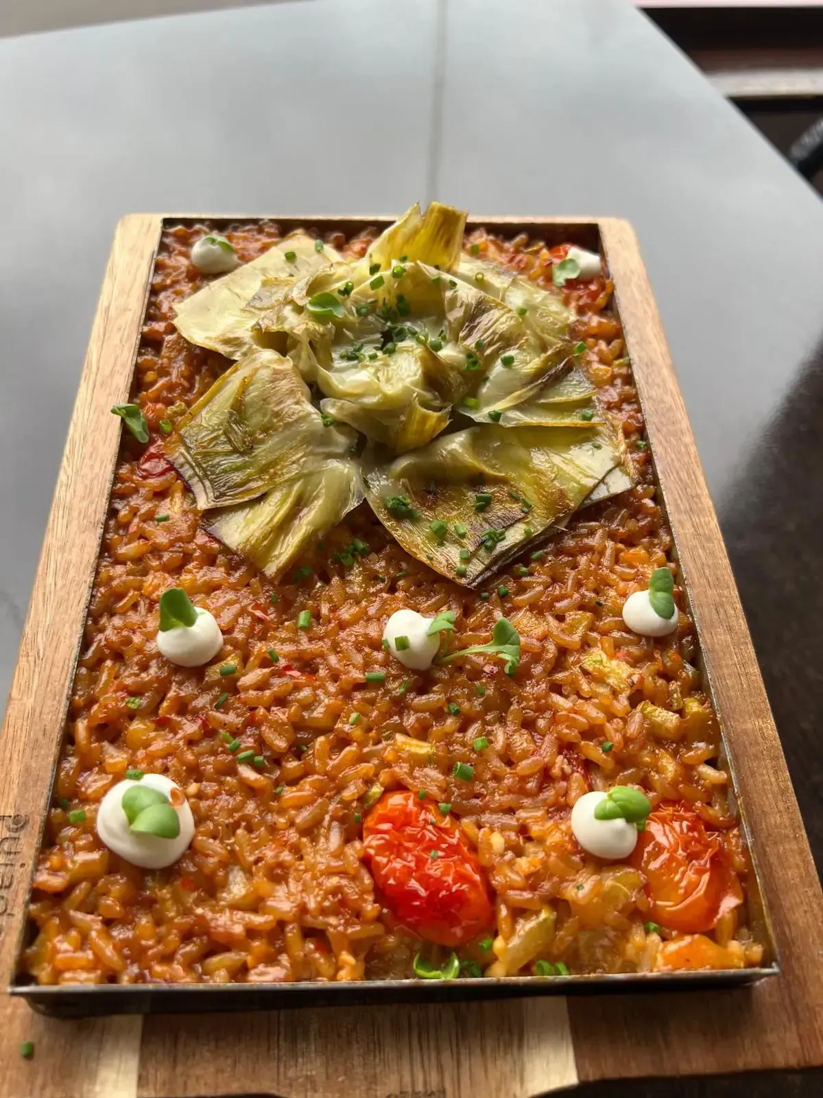 Una porción rectangular de risotto de color naranja, cubierto con corazones de alcachofa, tomates asados