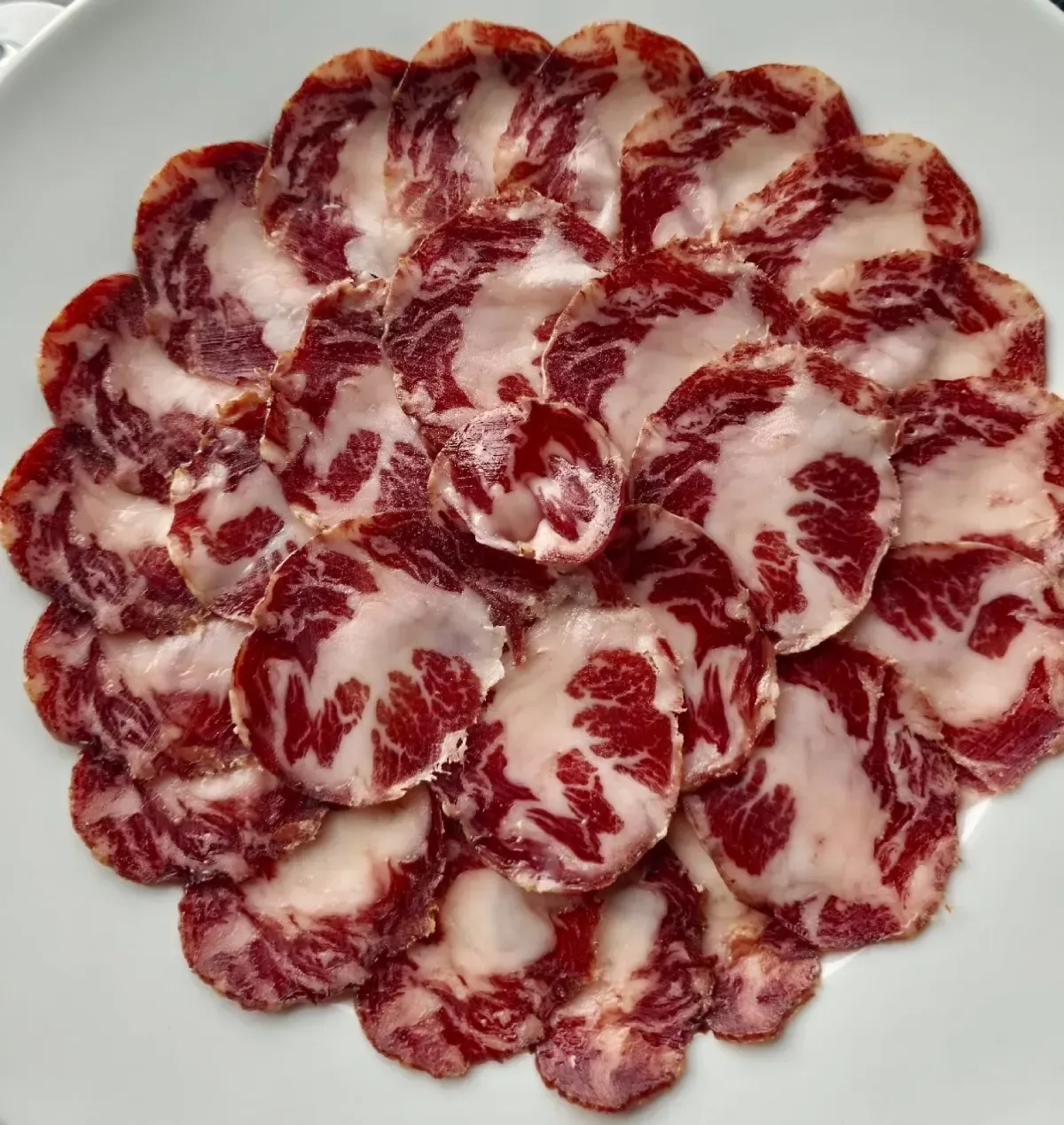 Carne curada veteada, cortada en finas lonchas, dispuesta en forma circular sobre un plato blanco.