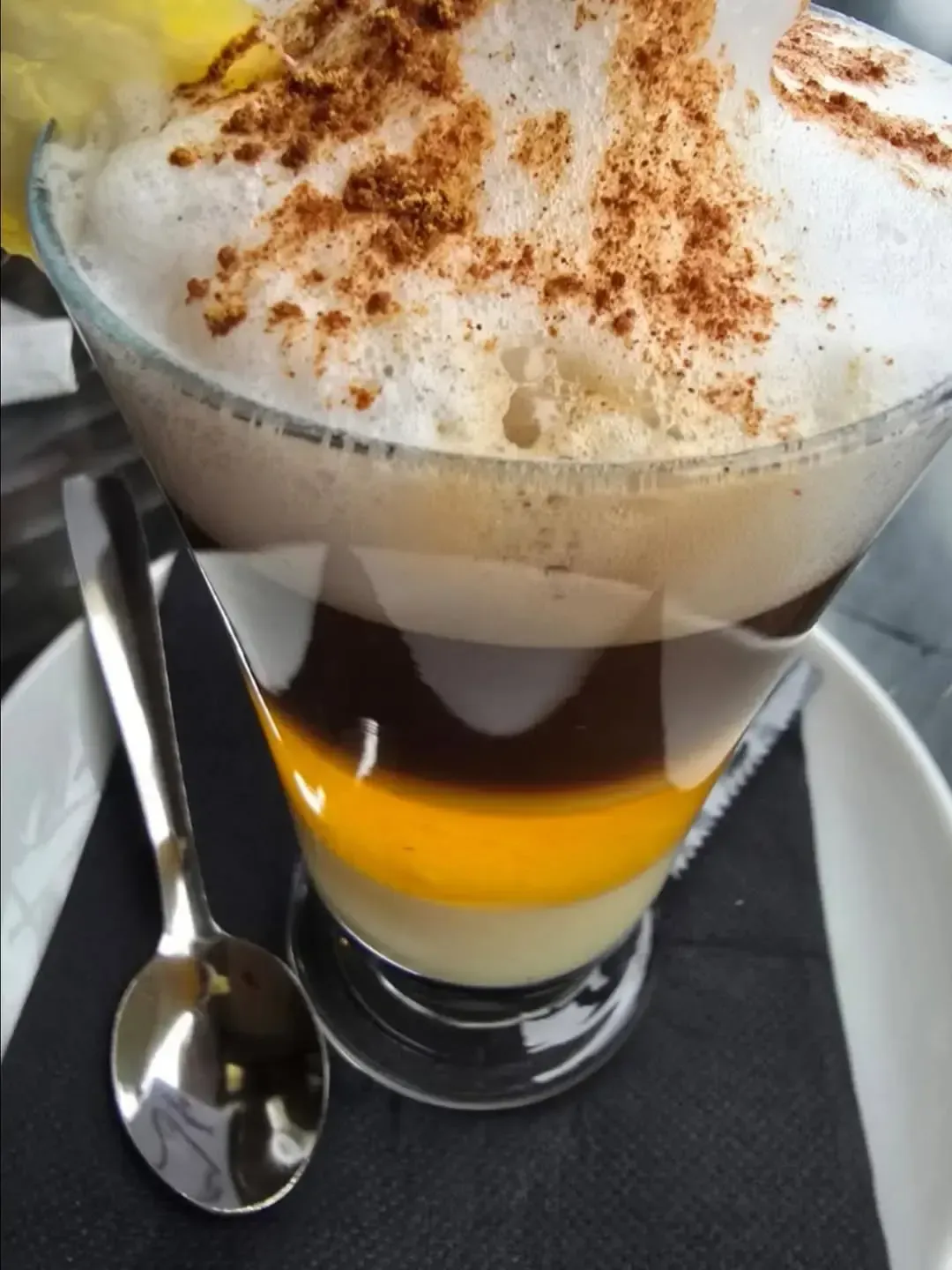 Un vaso alto de café en capas con una base amarilla, espresso oscuro, espuma de leche y una pizca de canela
