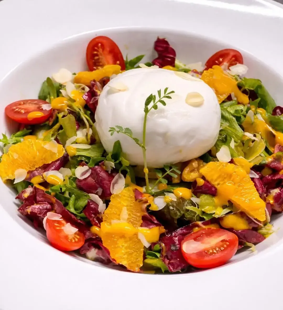 Una ensalada de burrata con tomates cherry, rodajas de naranja y hojas verdes en un bol blanco