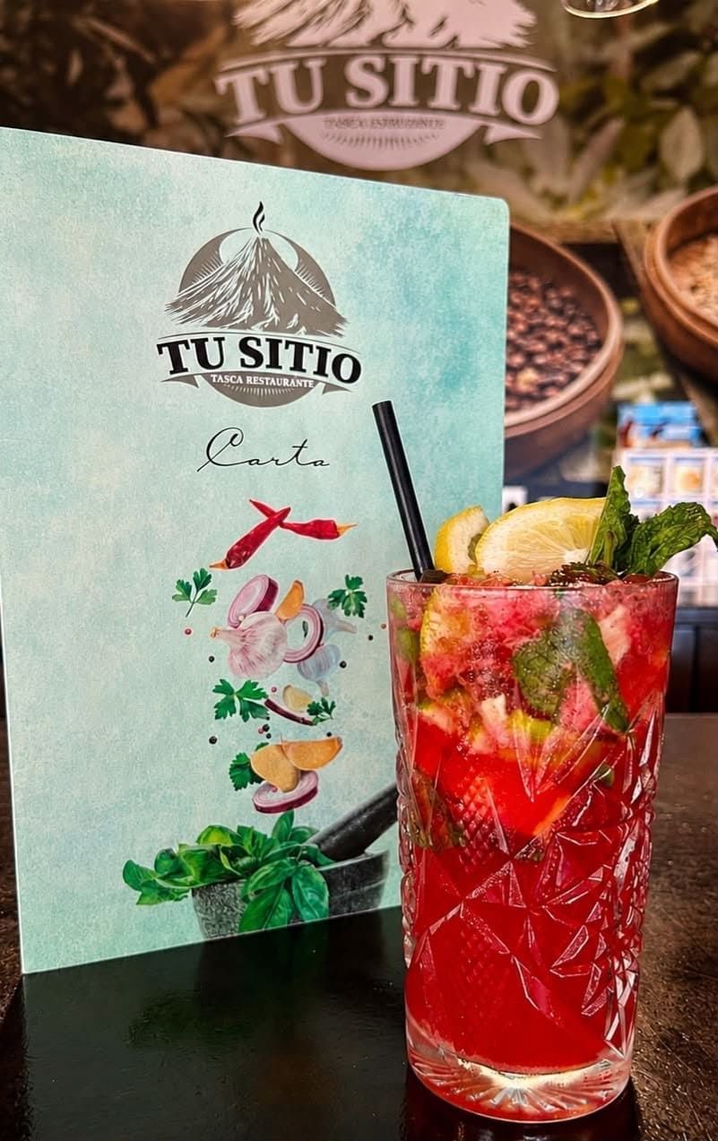 Un cóctel de frutas de color rojo brillante con lima sobre una mesa junto a un menú y un letrero de TU SITIO.