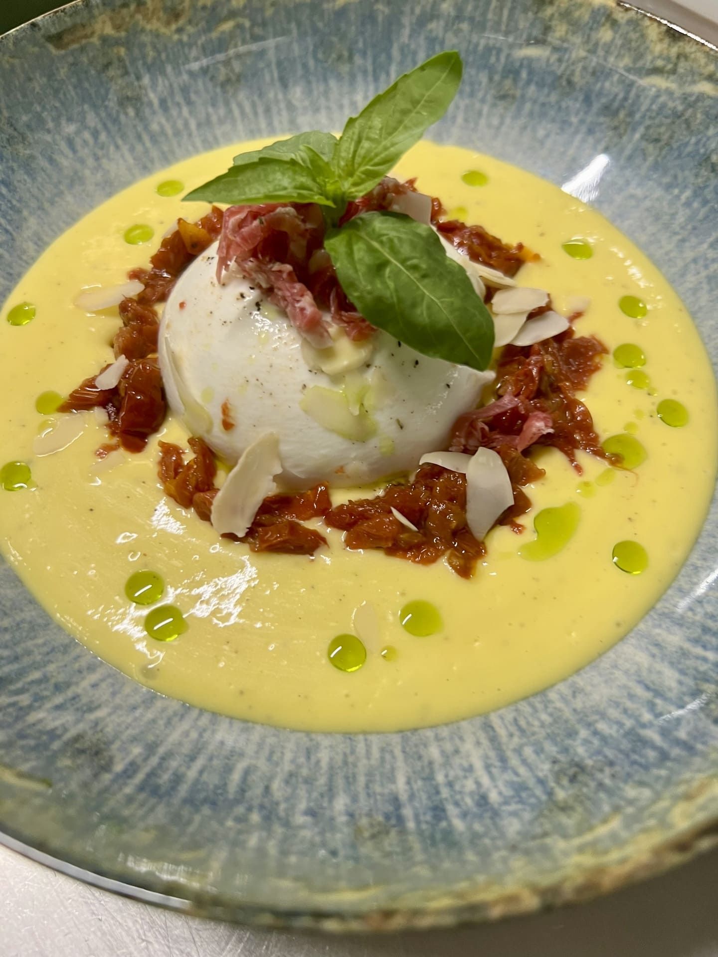 Postre helado en un tazón azul, cubierto con albahaca, nueces y salsa roja, con puntos verdes alrededor.