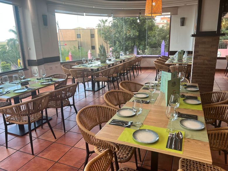 Vista interior de un restaurante vacío con sillas de mimbre negras, mesas cuadradas, suelos de baldosas 