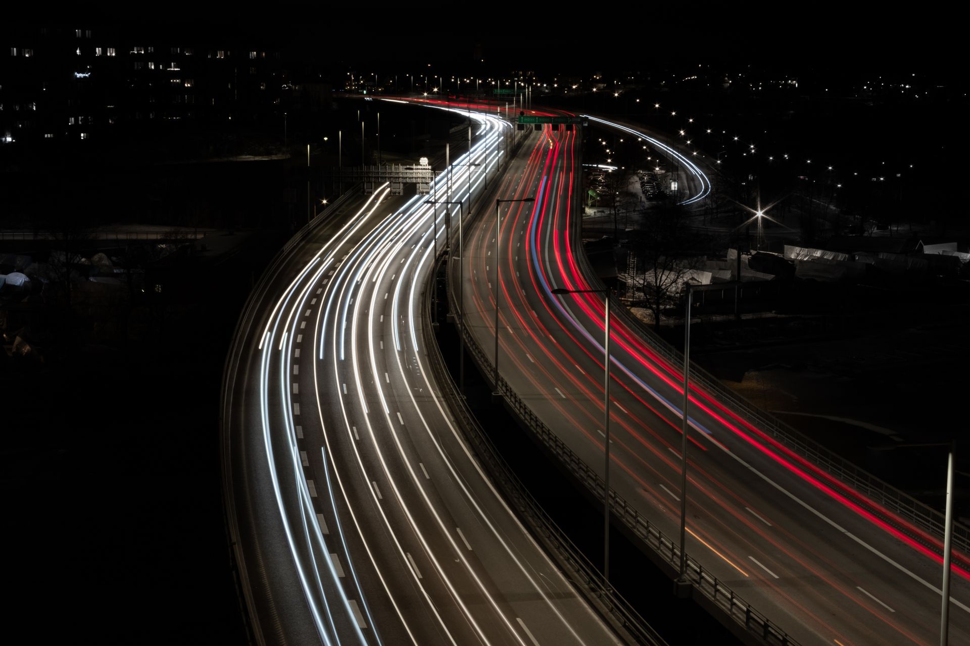 Autoroute de nuit, avec des traînées de phares blancs et de feux rouges qui serpentent à travers un paysage urbain sombre