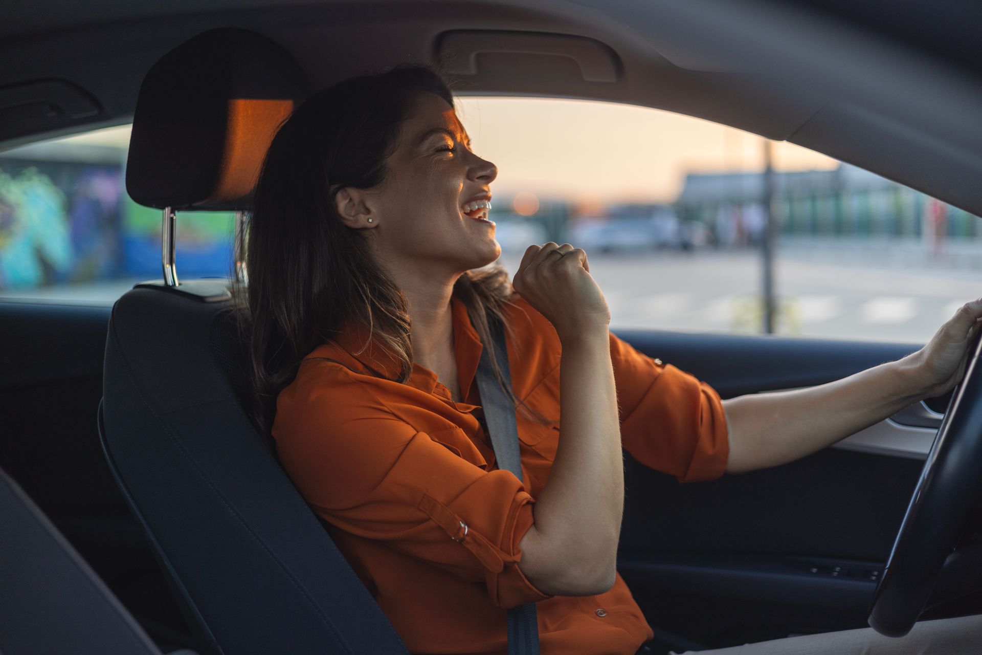 Une femme conduisant une voiture au coucher du soleil, vêtue d'un t-shirt orange et souriante, chantant en même temps.