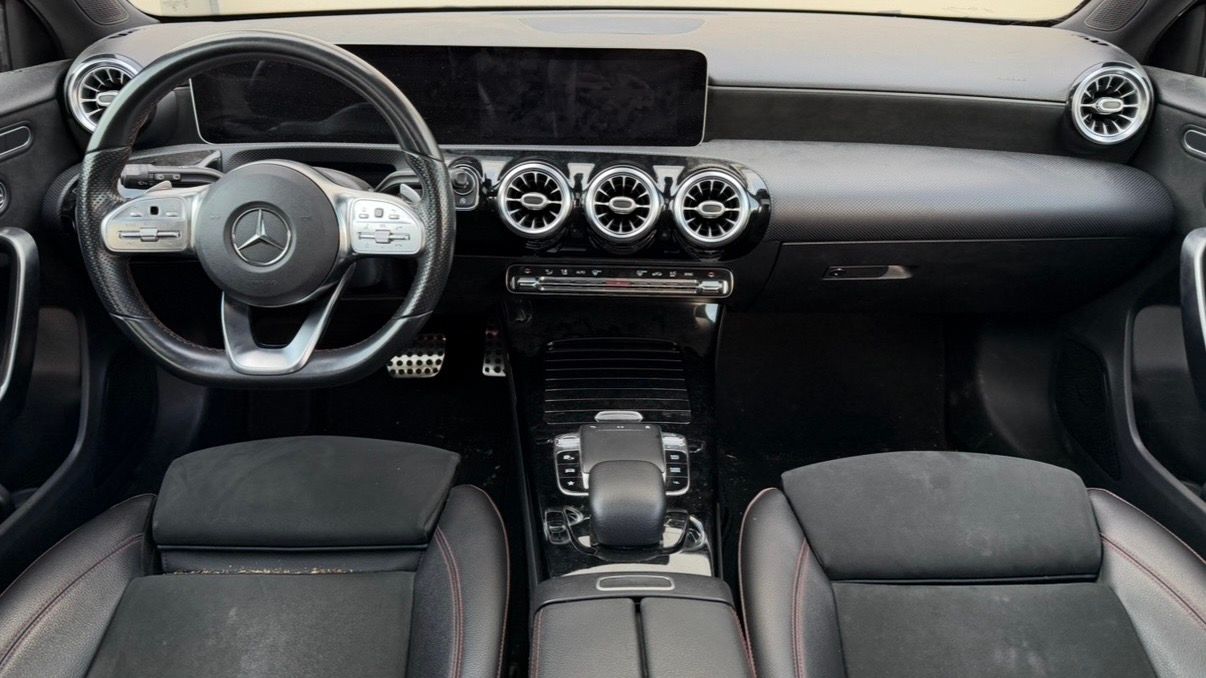 Intérieur moderne avec sièges noirs, volant Mercedes et aérateurs circulaires sur le tableau de bord sale.