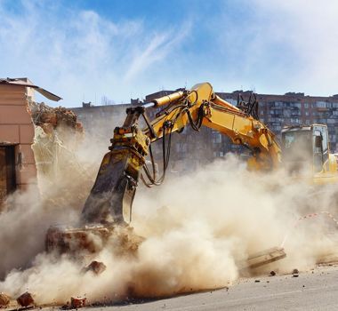 Una excavadora amarilla equipada con un martillo hidráulico demuele un edificio, levantando una gran nube de polvo.