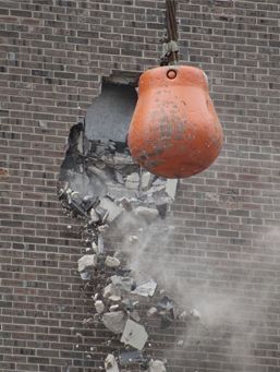 Una bola de demolición naranja se estrella contra una pared de ladrillos, provocando que caigan escombros y polvo.