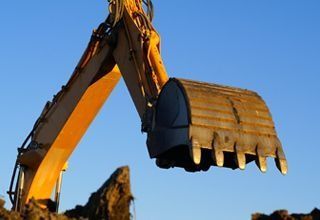 El brazo amarillo de una excavadora y su pala metálica se extienden sobre un montón de tierra oscura contra un cielo azul claro.