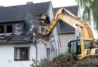Una excavadora amarilla demuele la parte superior del muro y el tejado de una casa.
