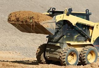 Una minicargadora amarilla transporta un cubo lleno de tierra a través de una obra en construcción arenosa.