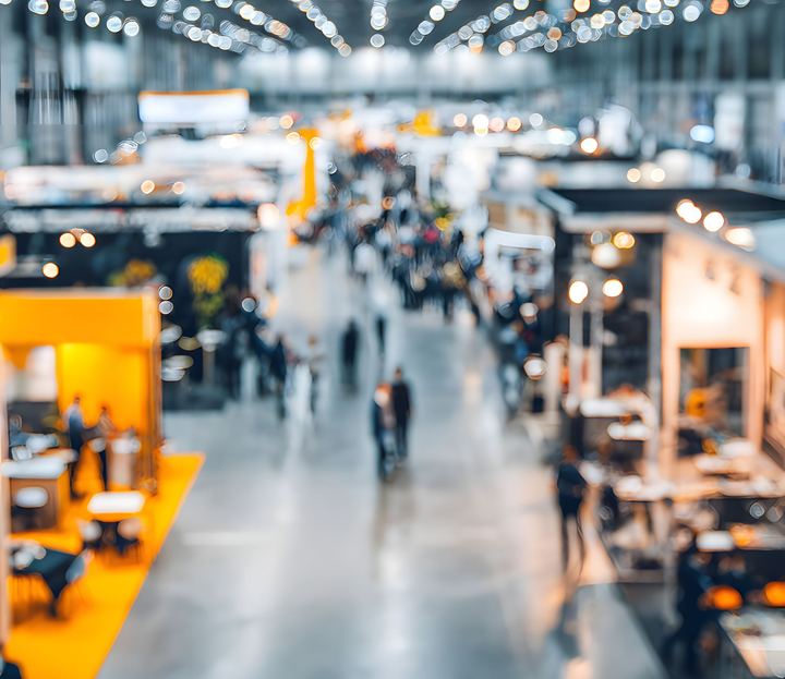 Vue aérienne floue d'une grande salle de congrès avec des stands, des personnes et des lumières vives au plafond.