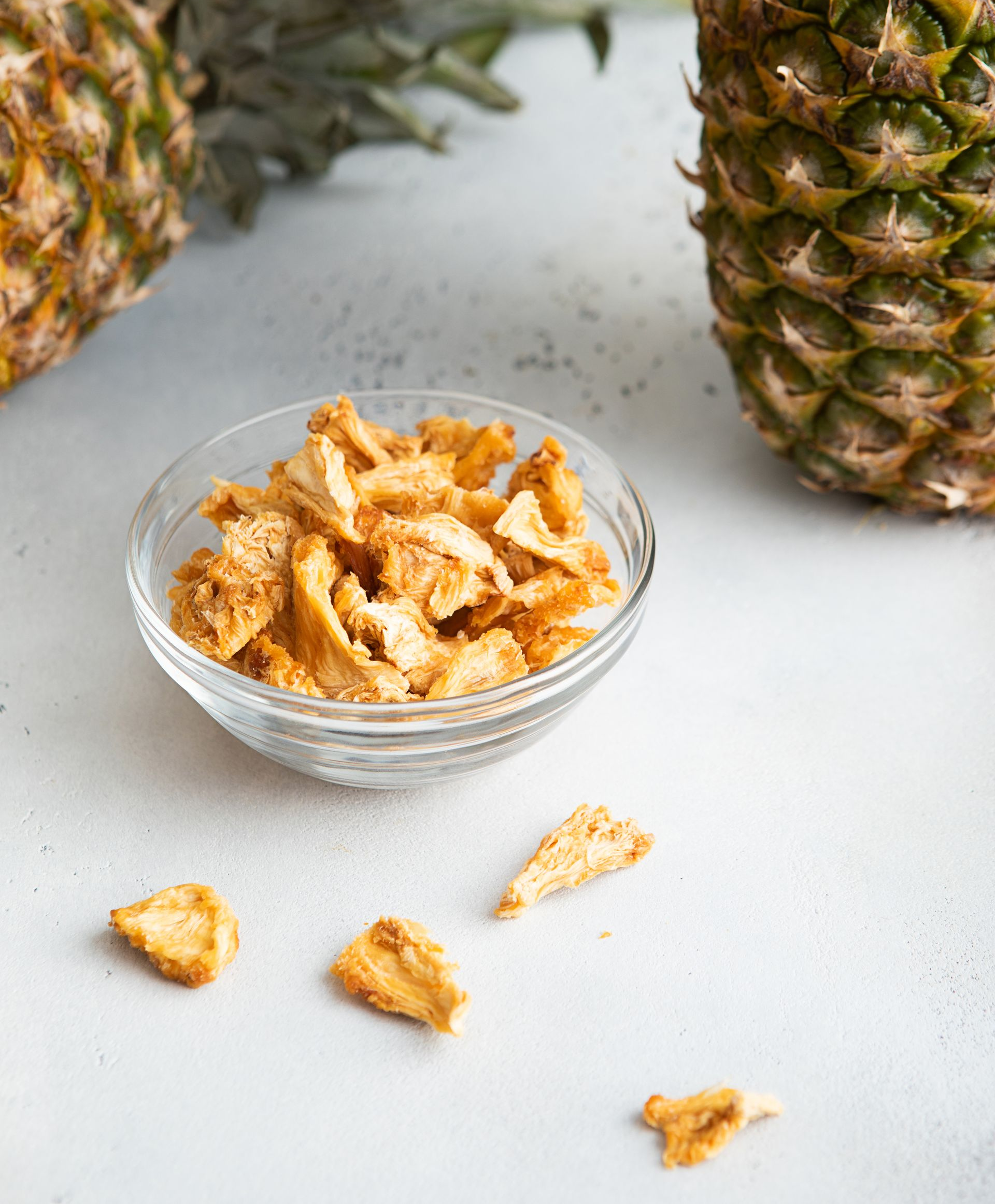 Bol de tranches d'ananas séchées avec deux ananas entiers sur une surface gris clair.