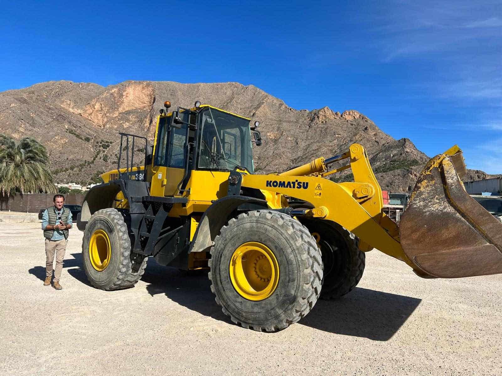 Hombre de pie junto a un gran cargador Komatsu amarillo con montañas al fondo.