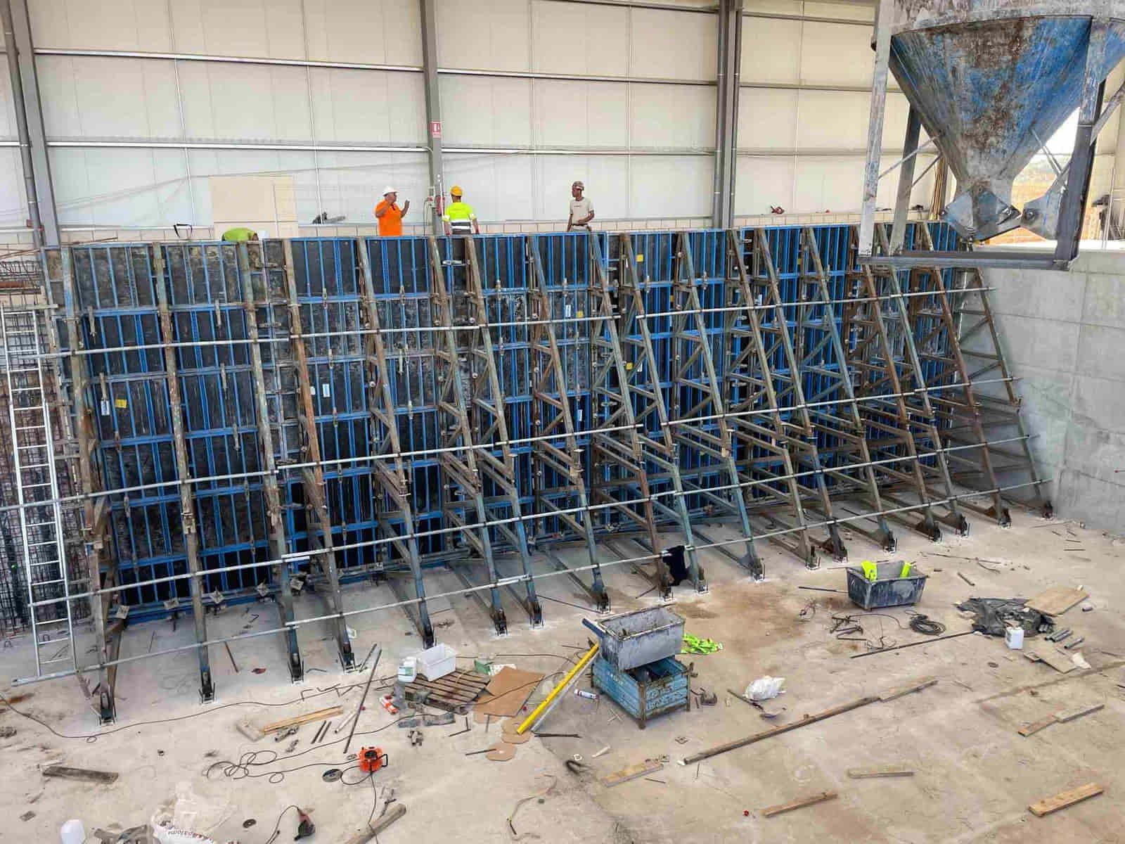 Obra: Obreros cerca de encofrados de hormigón azules, probablemente para un muro. Hormigón vertido al fondo.