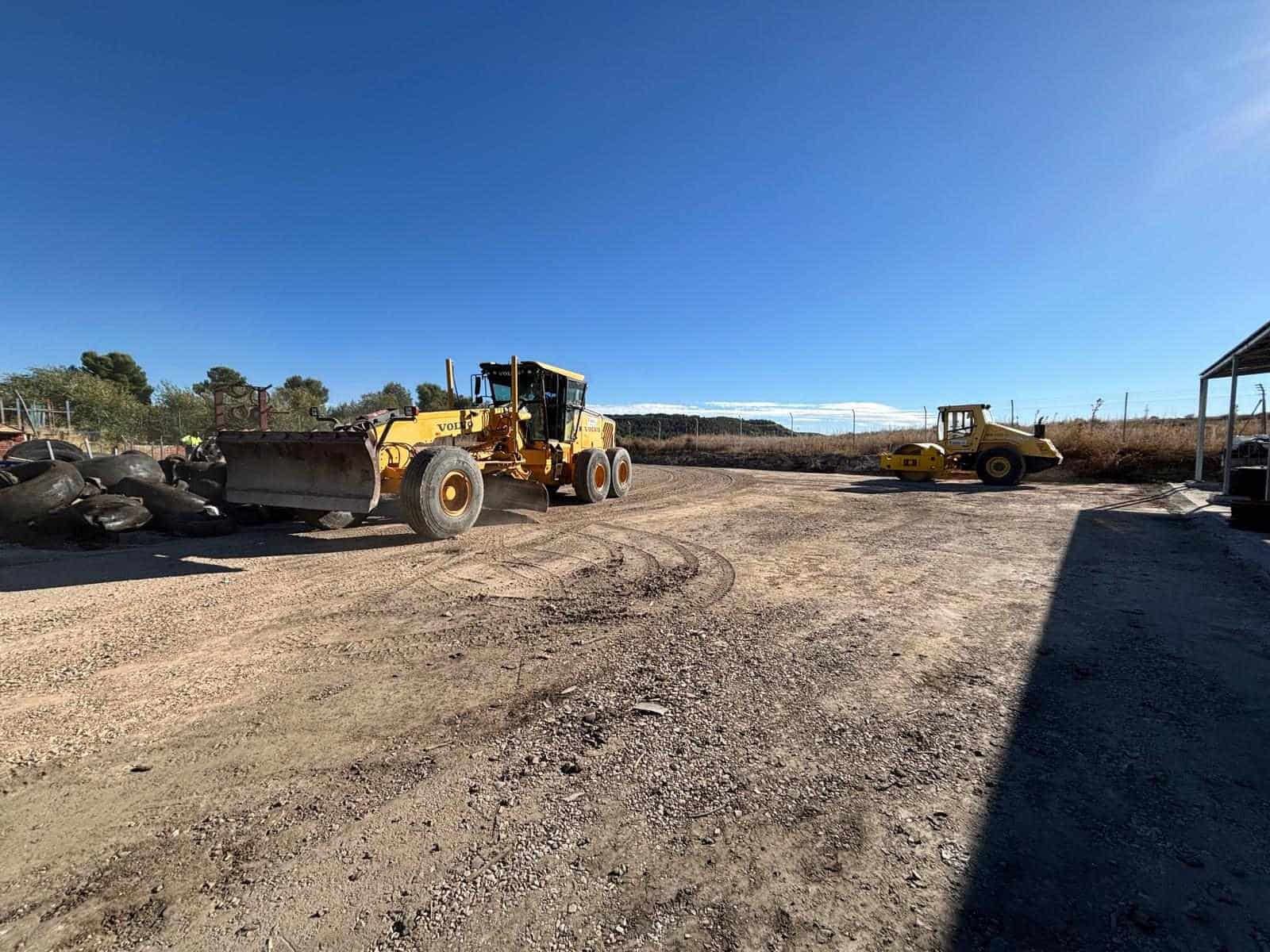 Una motoniveladora amarilla nivelando un terreno de grava con una excavadora estacionada cerca bajo un cielo azul.