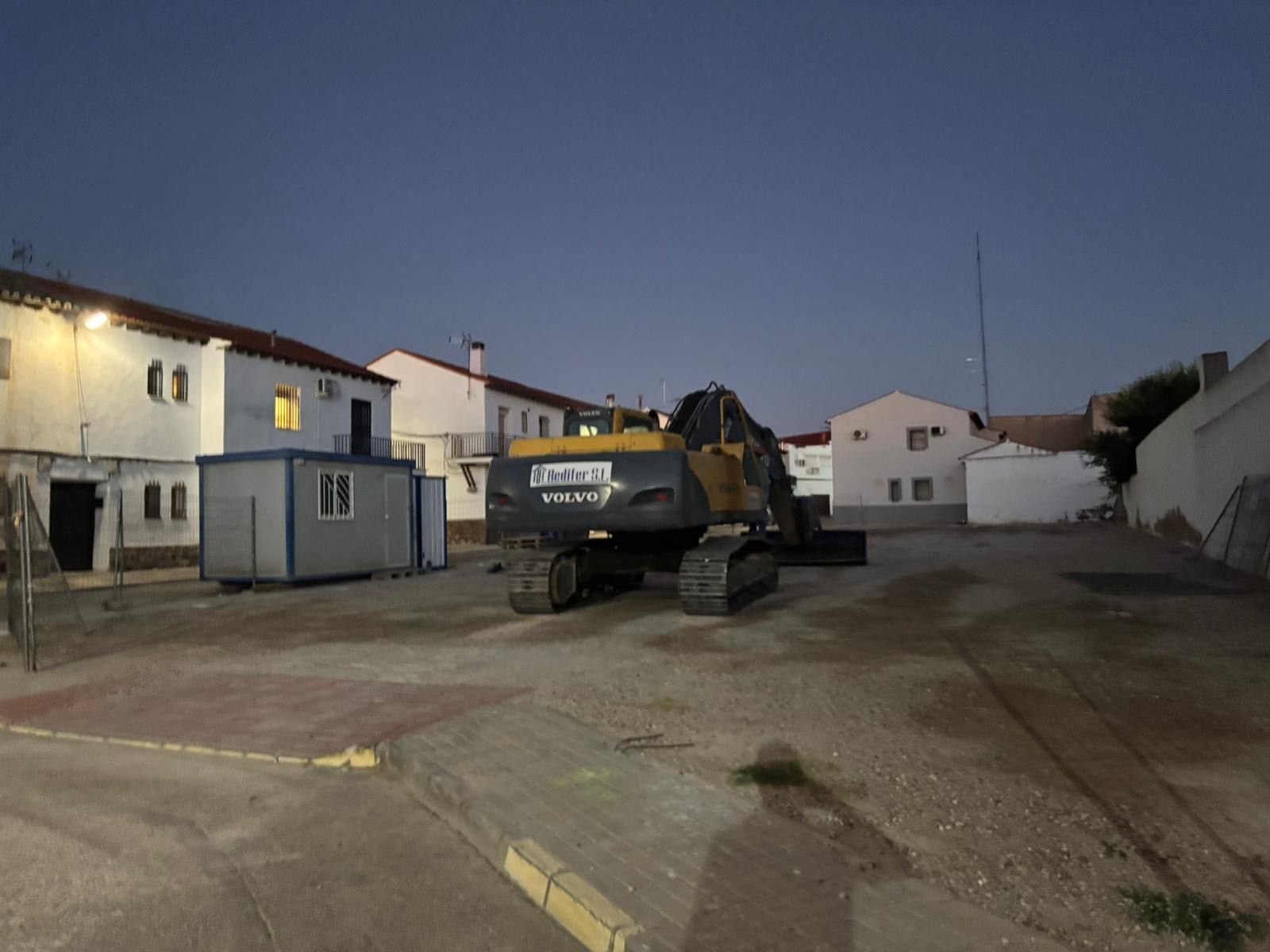 Excavadora estacionada en un lote de grava al anochecer, rodeada de edificios blancos.