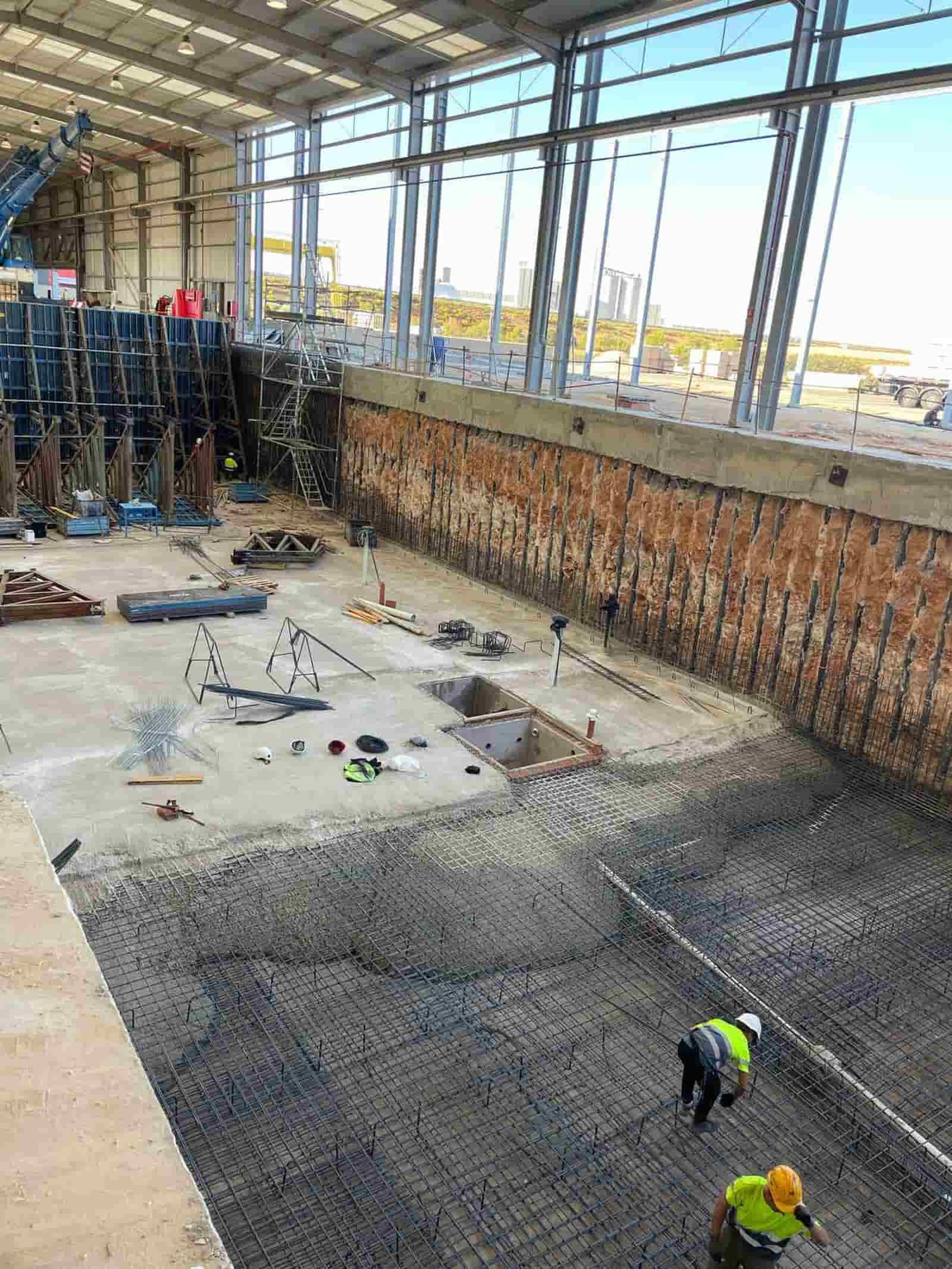 Obra: Obreros colocando varillas de refuerzo en un gran foso. Paredes marrones, cielo azul visible a través de las ventanas.