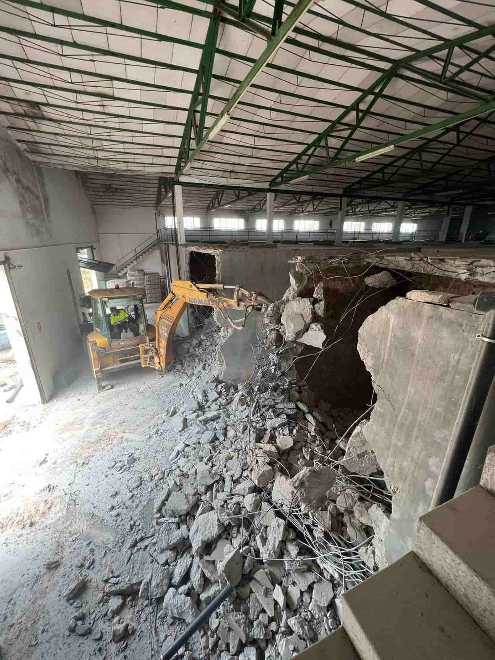 Una excavadora demuele una estructura de hormigón dentro de un gran edificio. Escena de construcción gris y amarilla.