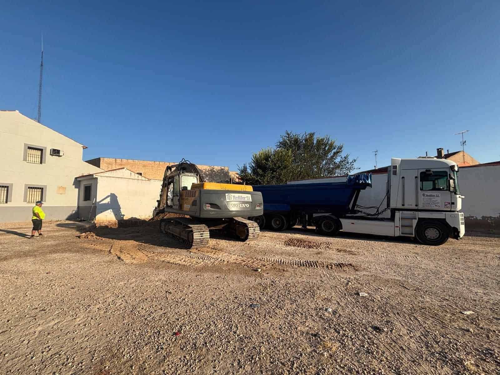 Obra con excavadora y camión volquete frente a un edificio. Se ve un trabajador.