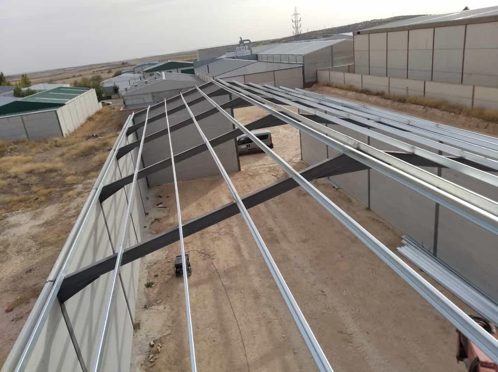 Construcción de estructura de techo de metal sobre un área de tierra, con edificios al fondo, entorno al aire libre.