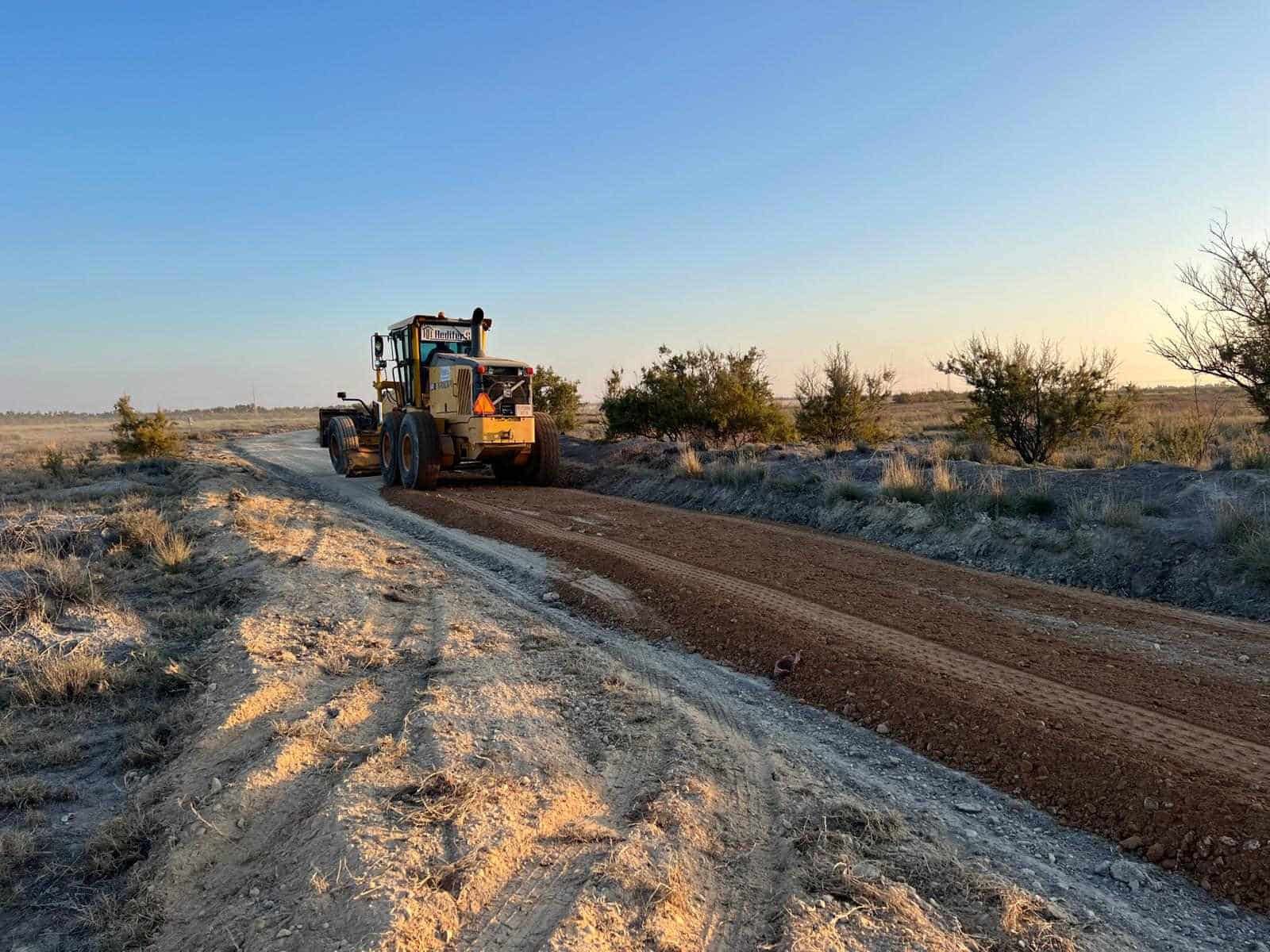 Una gran niveladora amarilla nivela un camino de tierra en un entorno rural bajo un cielo azul.