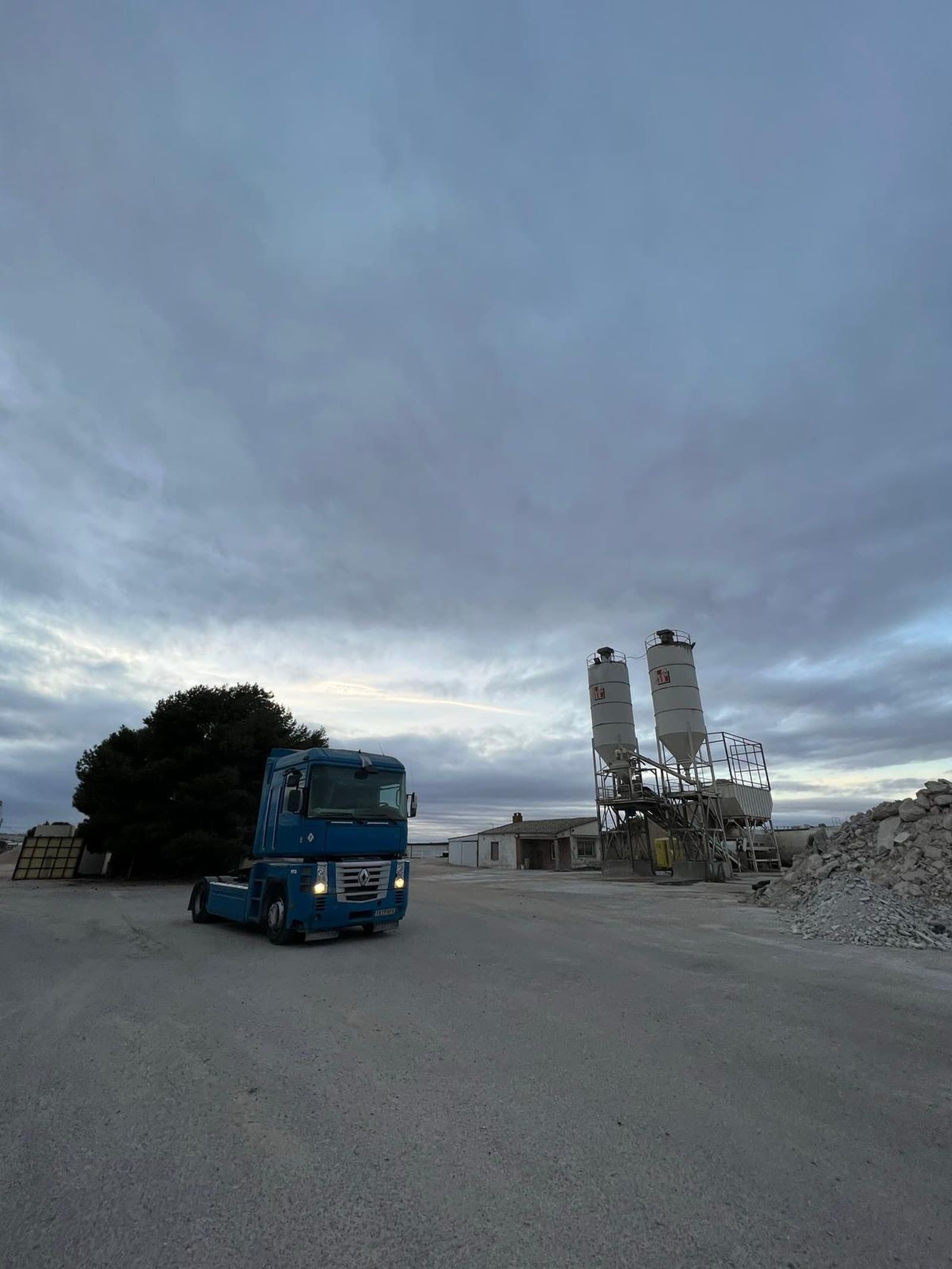 Camión azul estacionado frente a una planta de concreto bajo un cielo nublado.