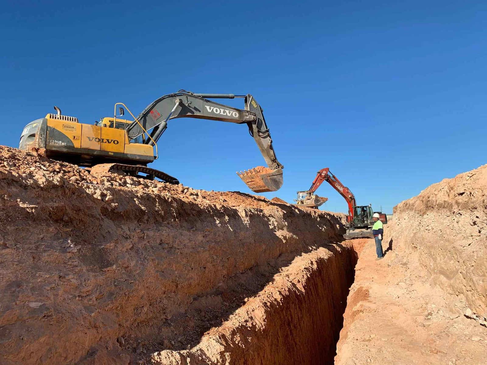 Dos excavadoras cavan una zanja profunda en un campo abierto de tierra de color marrón rojizo.