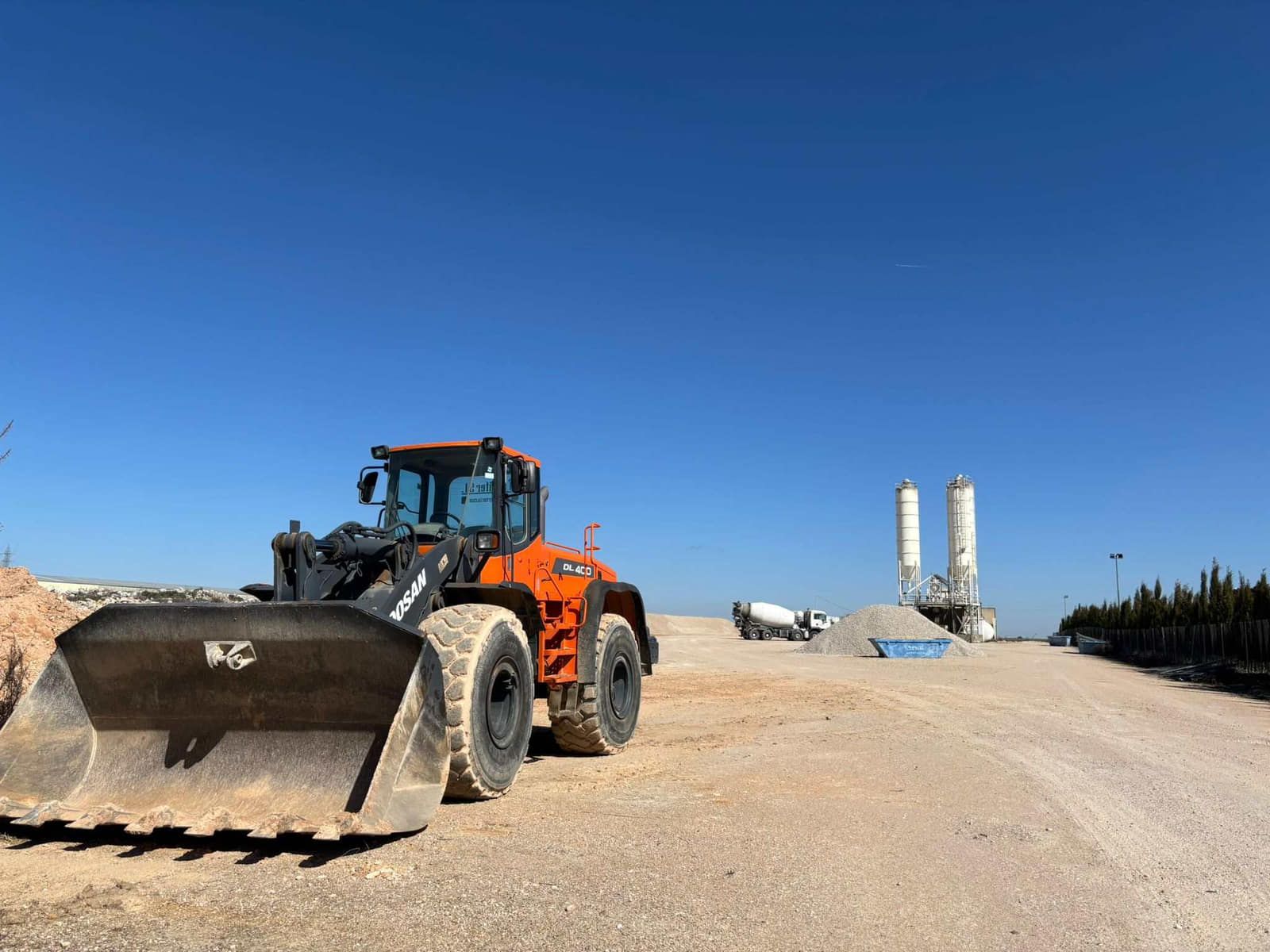 Cargador naranja en un camino de grava en un sitio de construcción con cielo azul y estructuras de cemento.