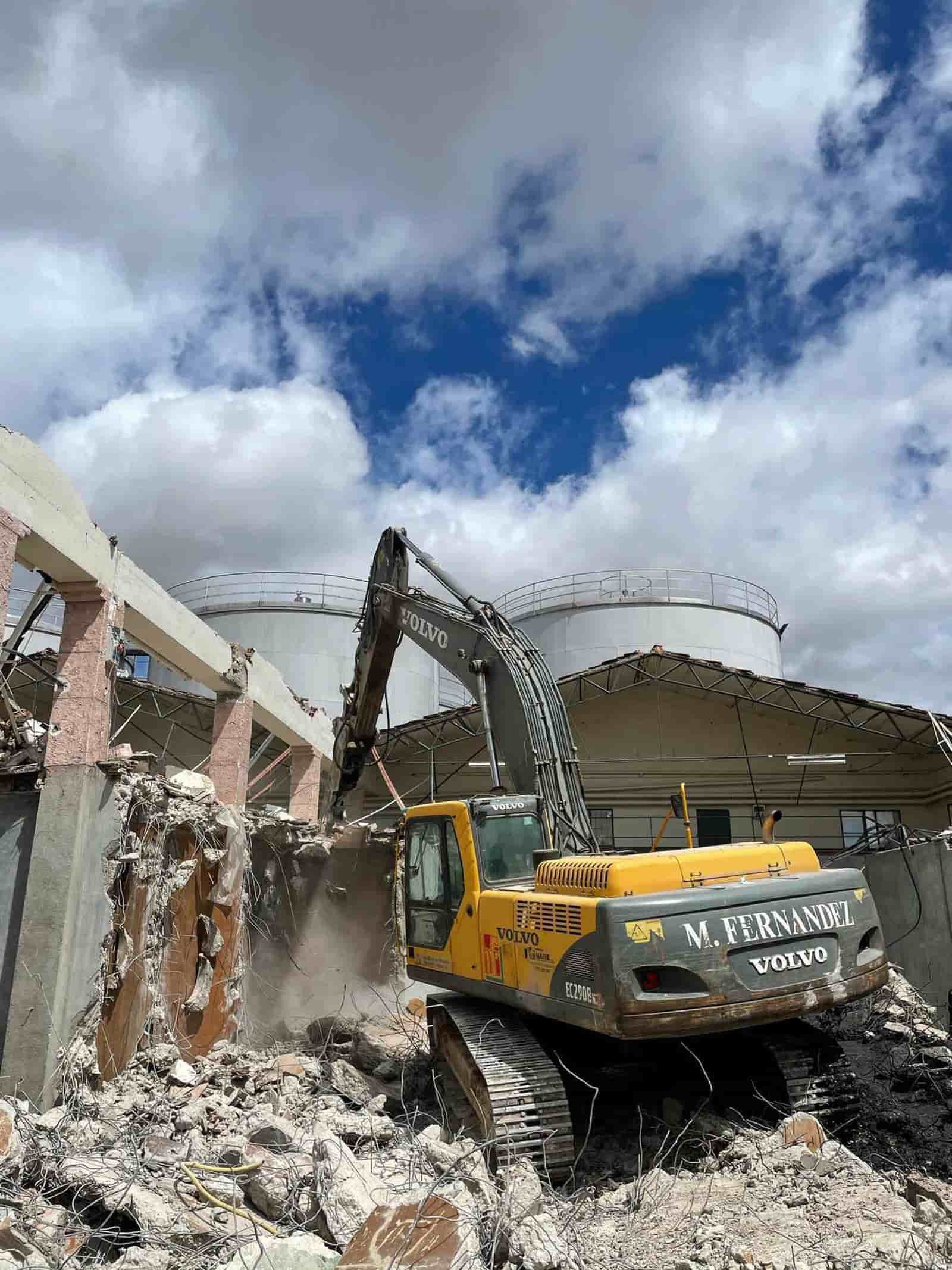 Una excavadora amarilla derriba un edificio bajo un cielo nublado, con tanques blancos en el fondo.