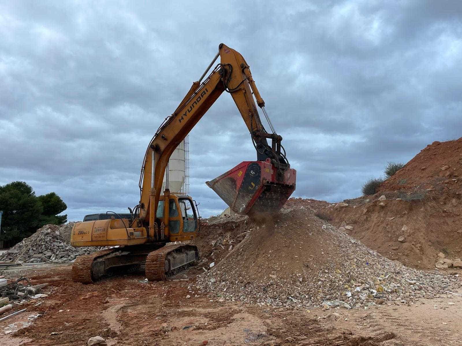 Excavadora amarilla cargando escombros de una pila bajo un cielo nublado.