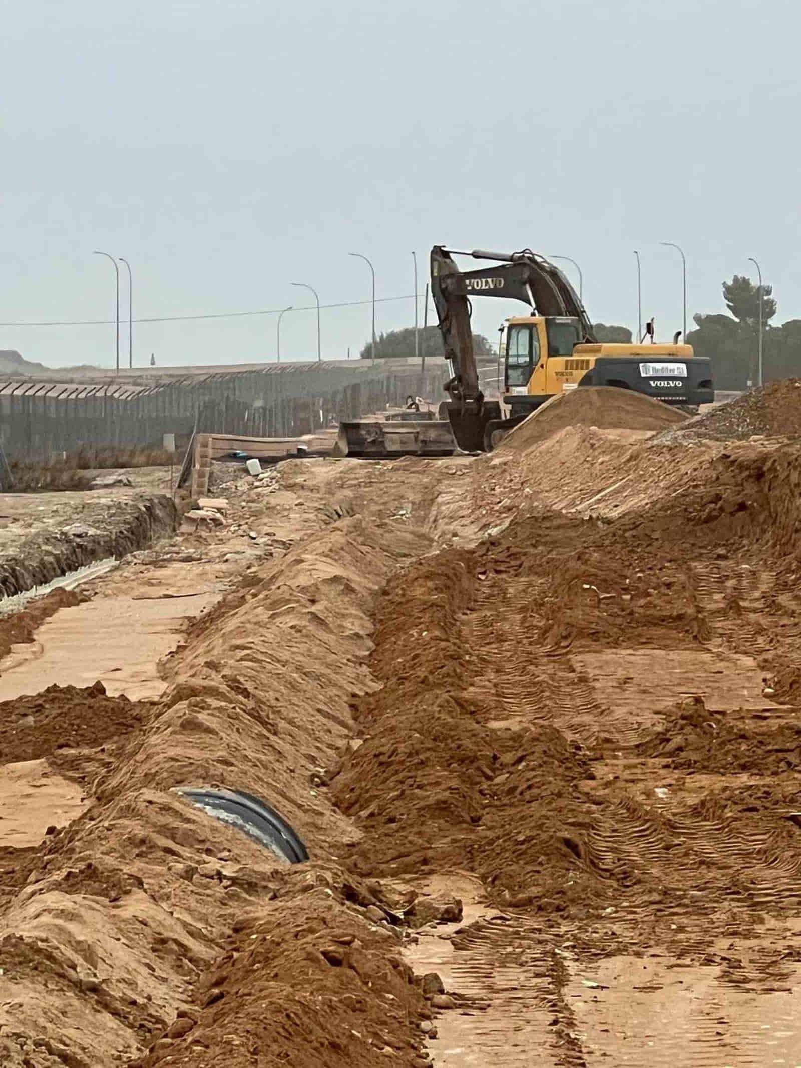 Una excavadora excava en un sitio de construcción, montones de tierra, puente al fondo.