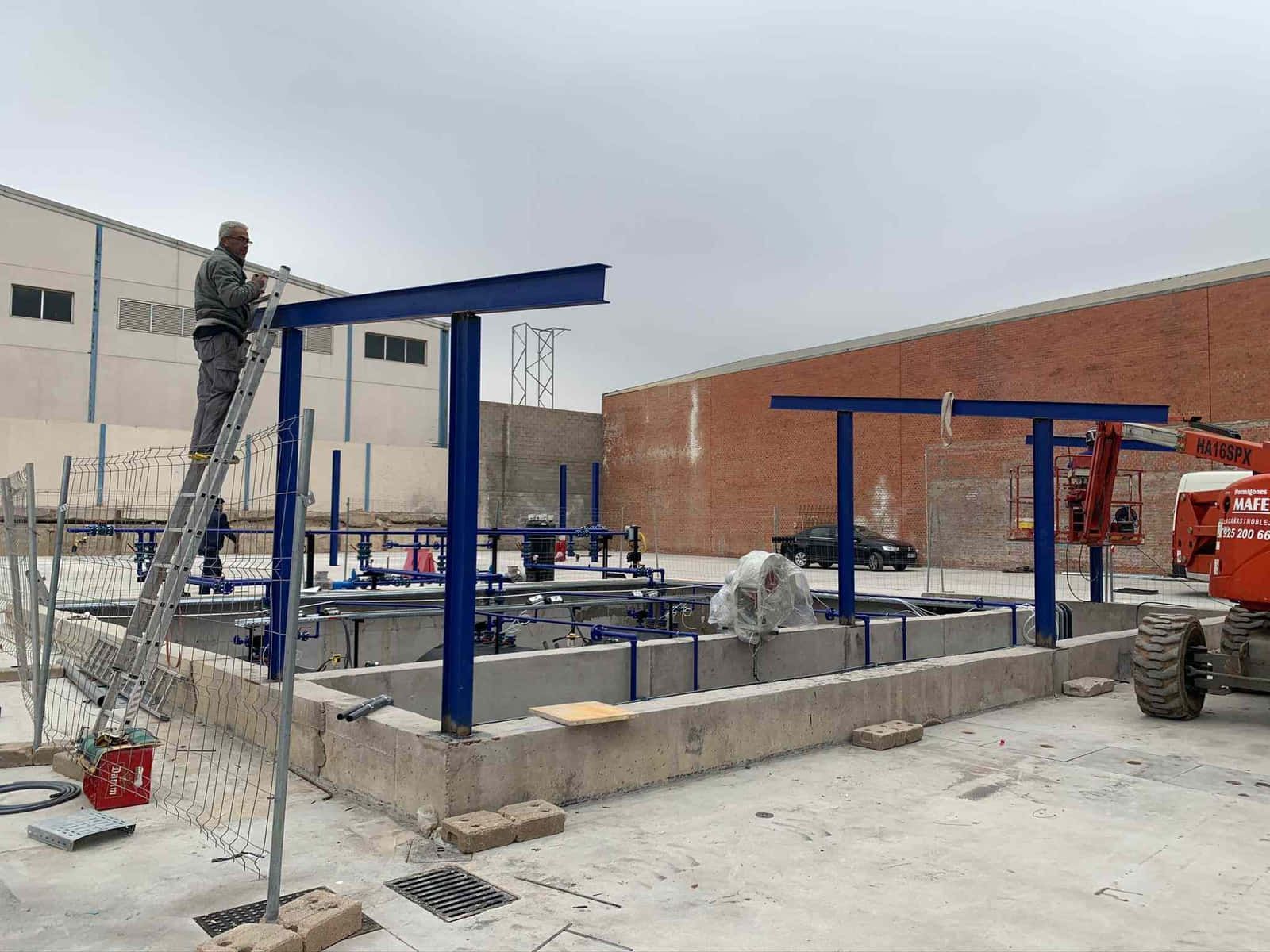 Obrero de la construcción en una escalera instalando vigas de metal azules sobre cuencas de hormigón.