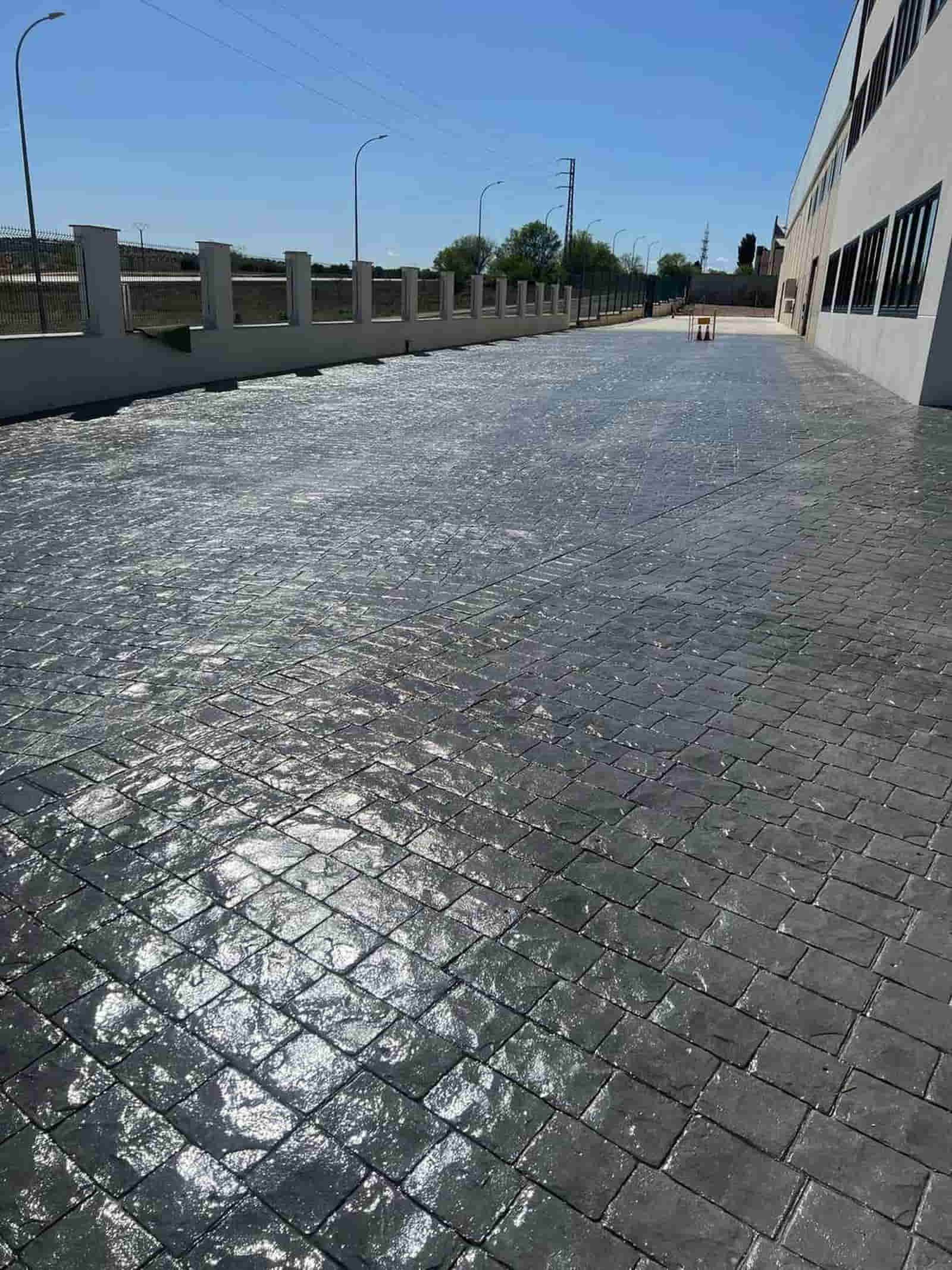 Superficie de hormigón con diseño de ladrillo que refleja la luz del sol junto a un edificio y una valla en un día soleado.