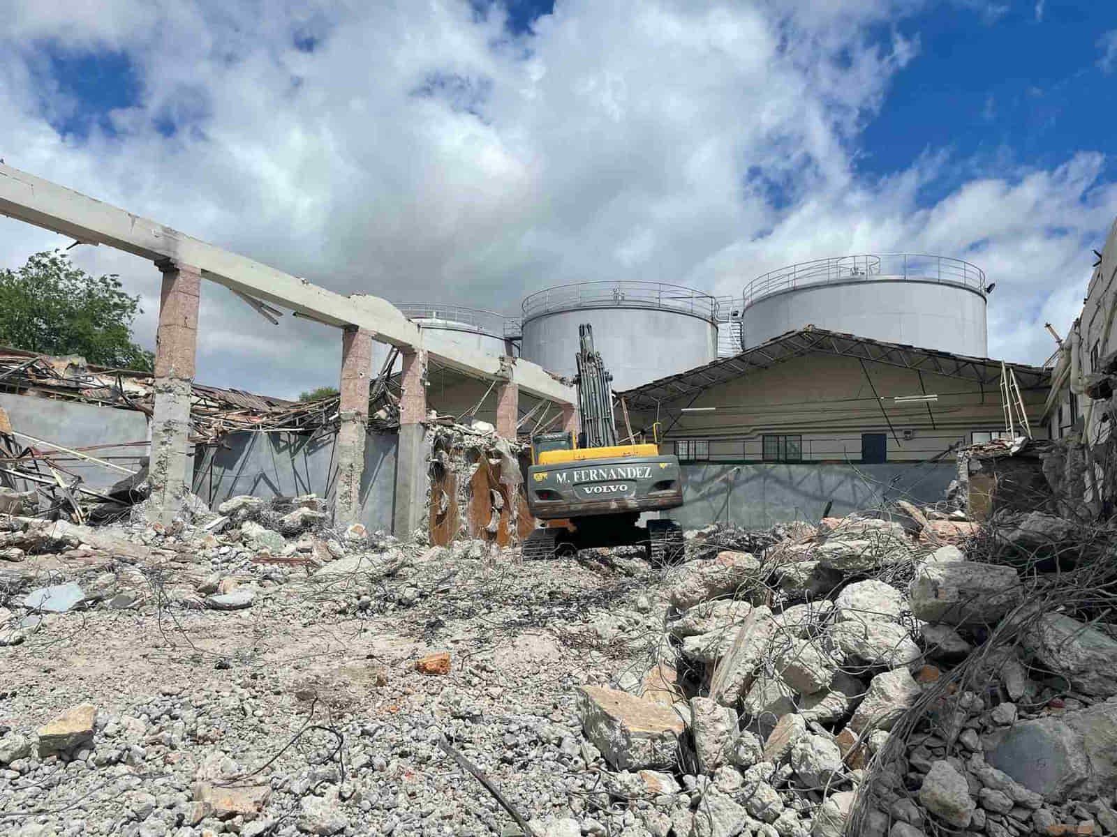 Una excavadora demuele un edificio, dejando escombros esparcidos. Al fondo se ven grandes tanques y un cielo nublado.