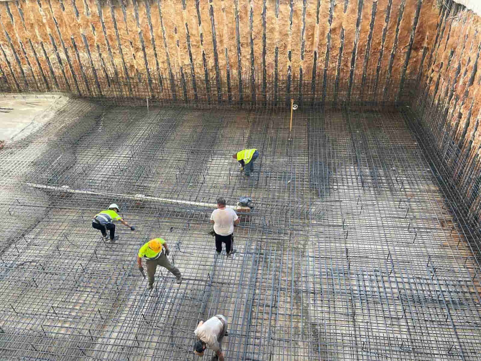 Obreros instalando varillas de refuerzo en una gran zanja excavada. Se ven chalecos de seguridad amarillos.