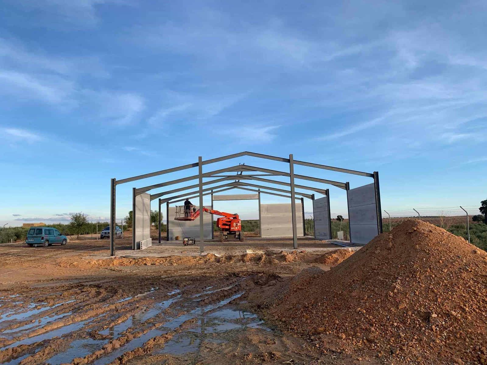 Edificio con estructura de acero en construcción en un campo fangoso con trabajadores en un ascensor.