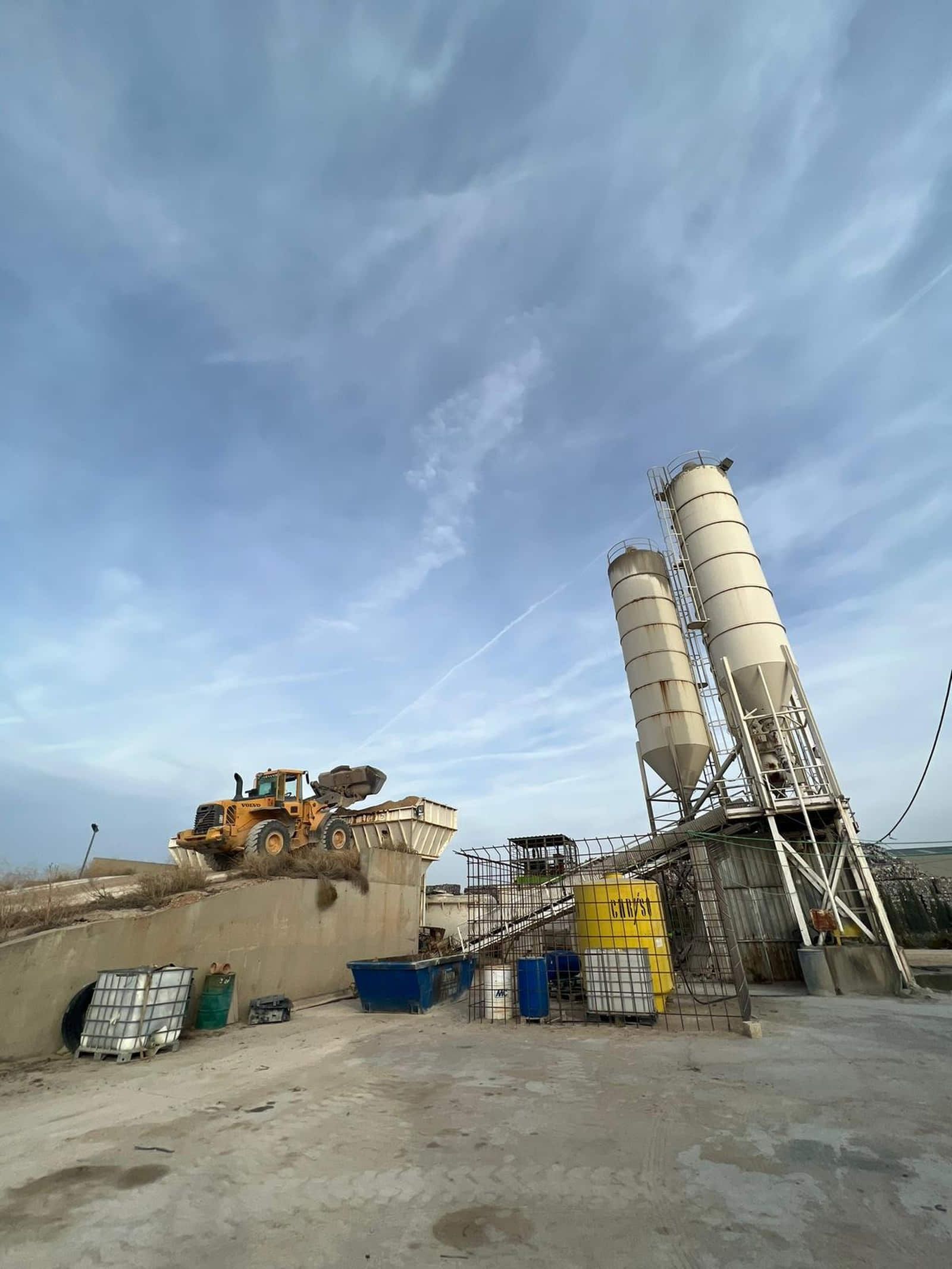 Planta de hormigón con silos, cargador y equipos bajo un cielo azul nublado.