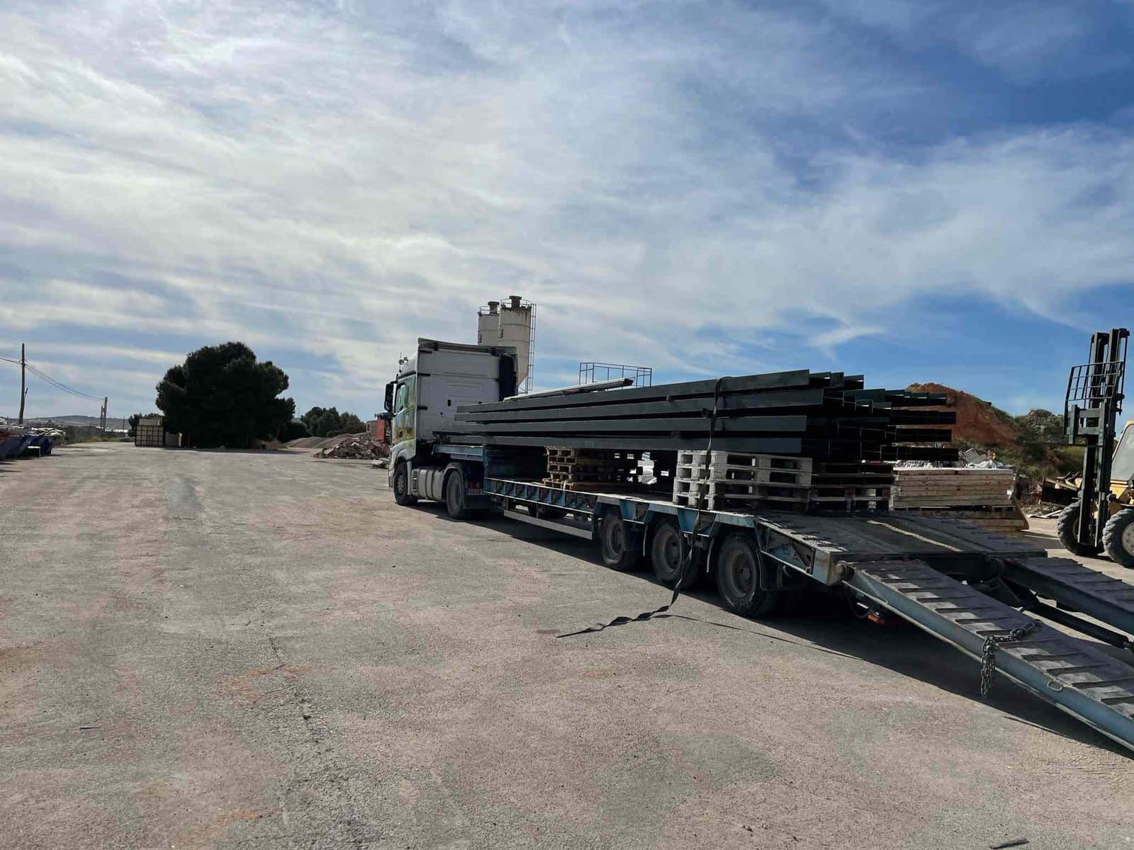 Camión semirremolque con remolque de plataforma larga que transporta vigas de metal apiladas en un lote de grava.