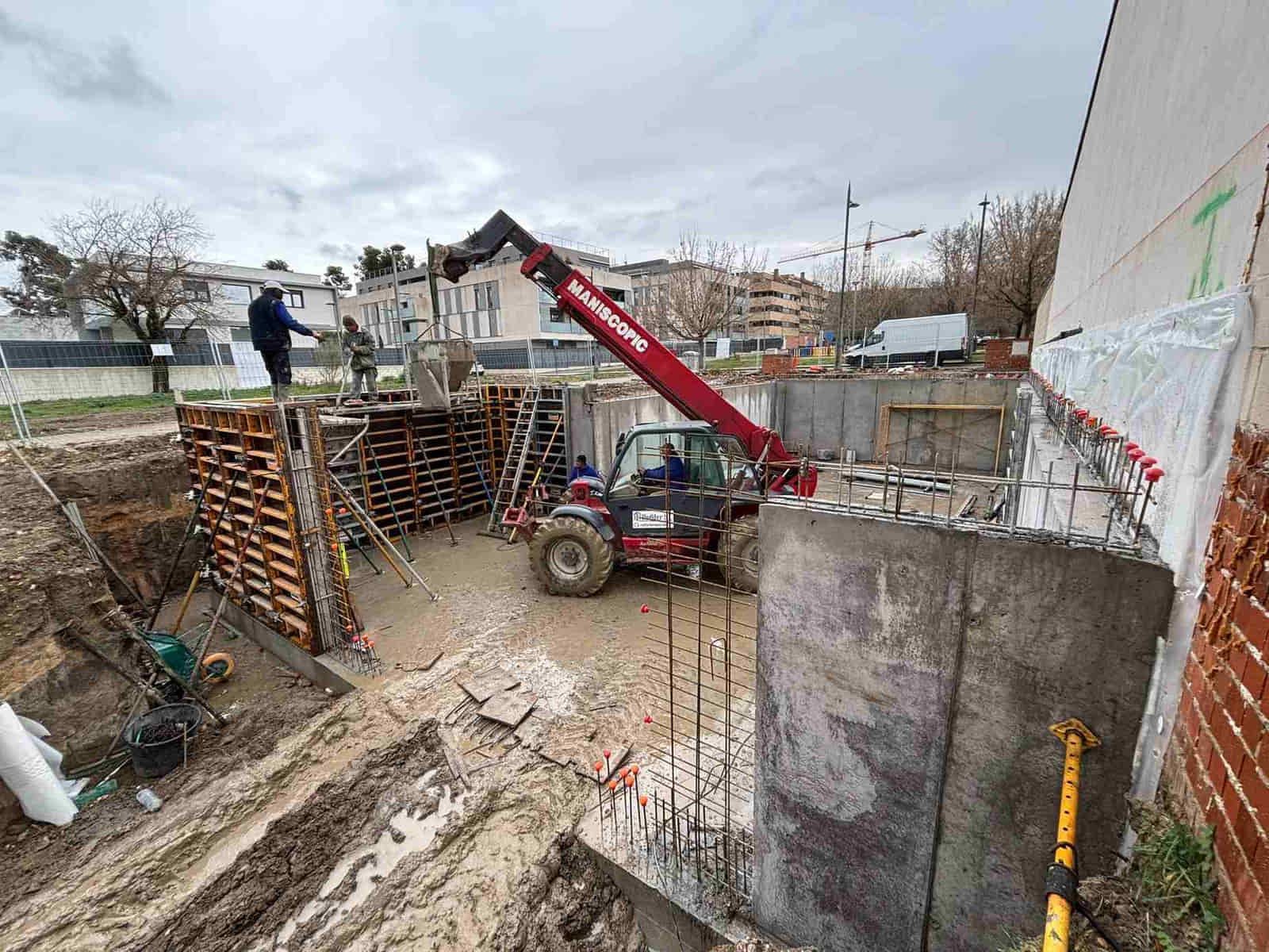 Obra con manipulador telescópico rojo y trabajadores. Zanja fangosa, muros de hormigón y soportes de madera.