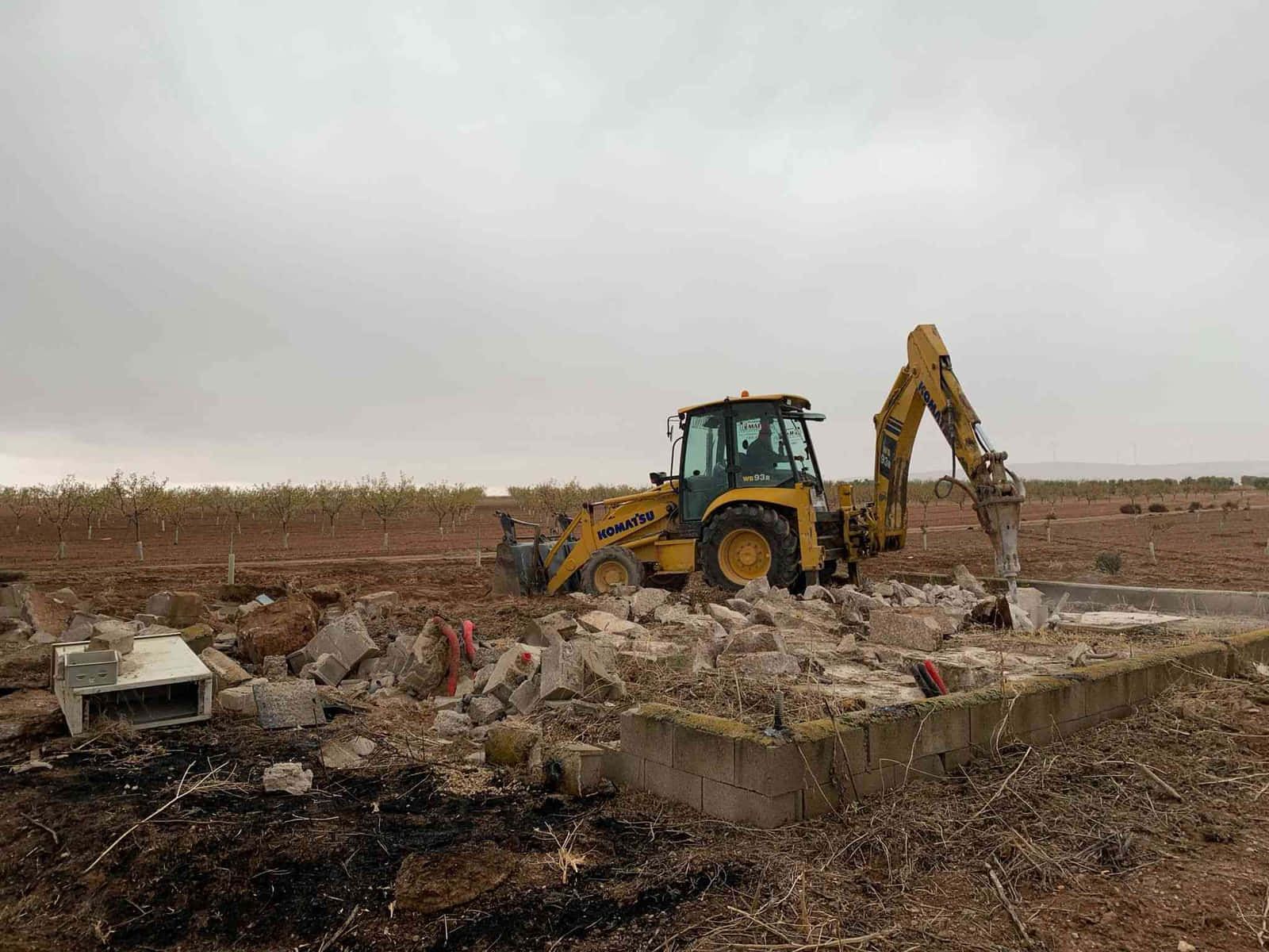 Una retroexcavadora amarilla derriba una estructura de hormigón en un campo bajo un cielo nublado.