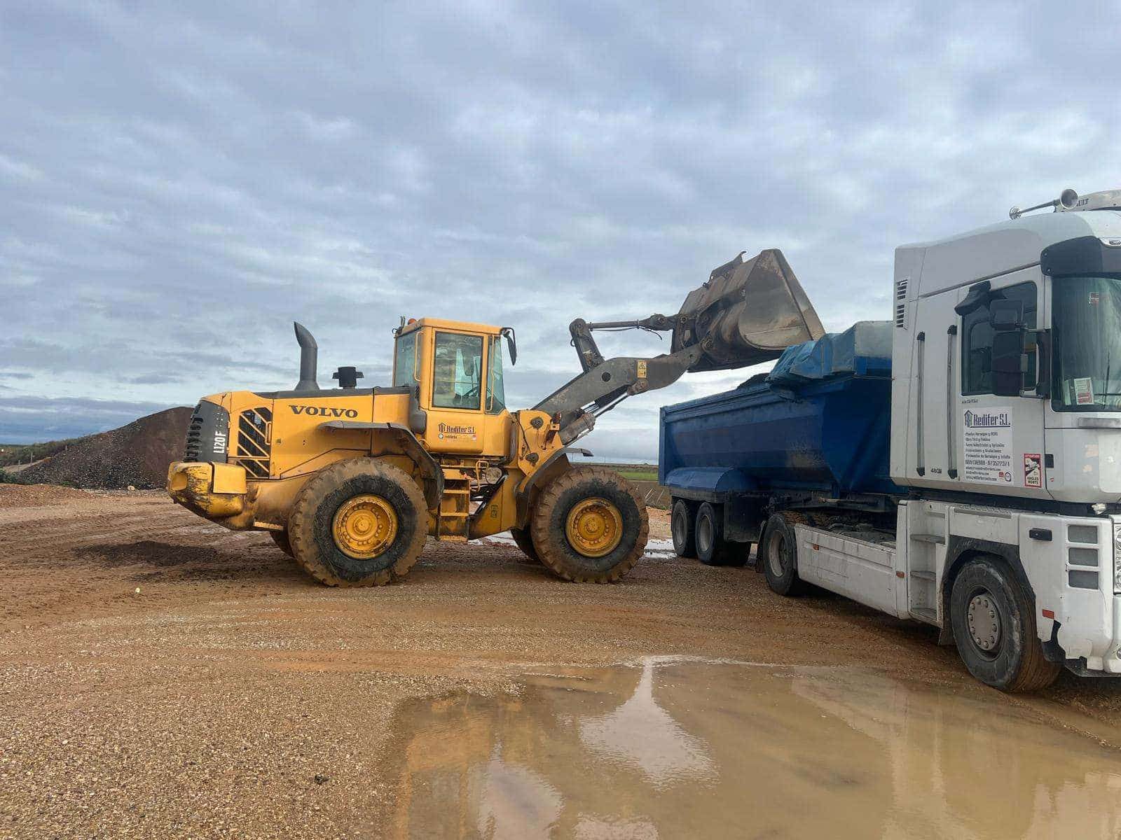 Un cargador amarillo llena un camión volquete azul con grava en un sitio de construcción bajo un cielo nublado.