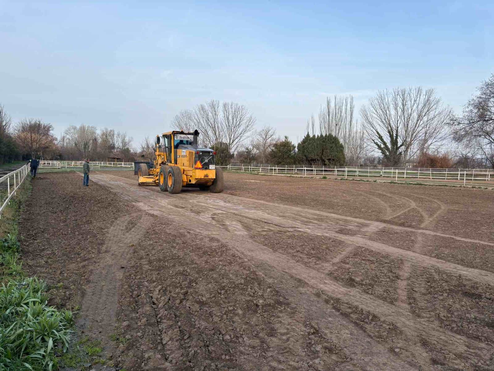 Una niveladora amarilla nivela una pista de tierra, con dos personas observando, bajo un cielo nublado.
