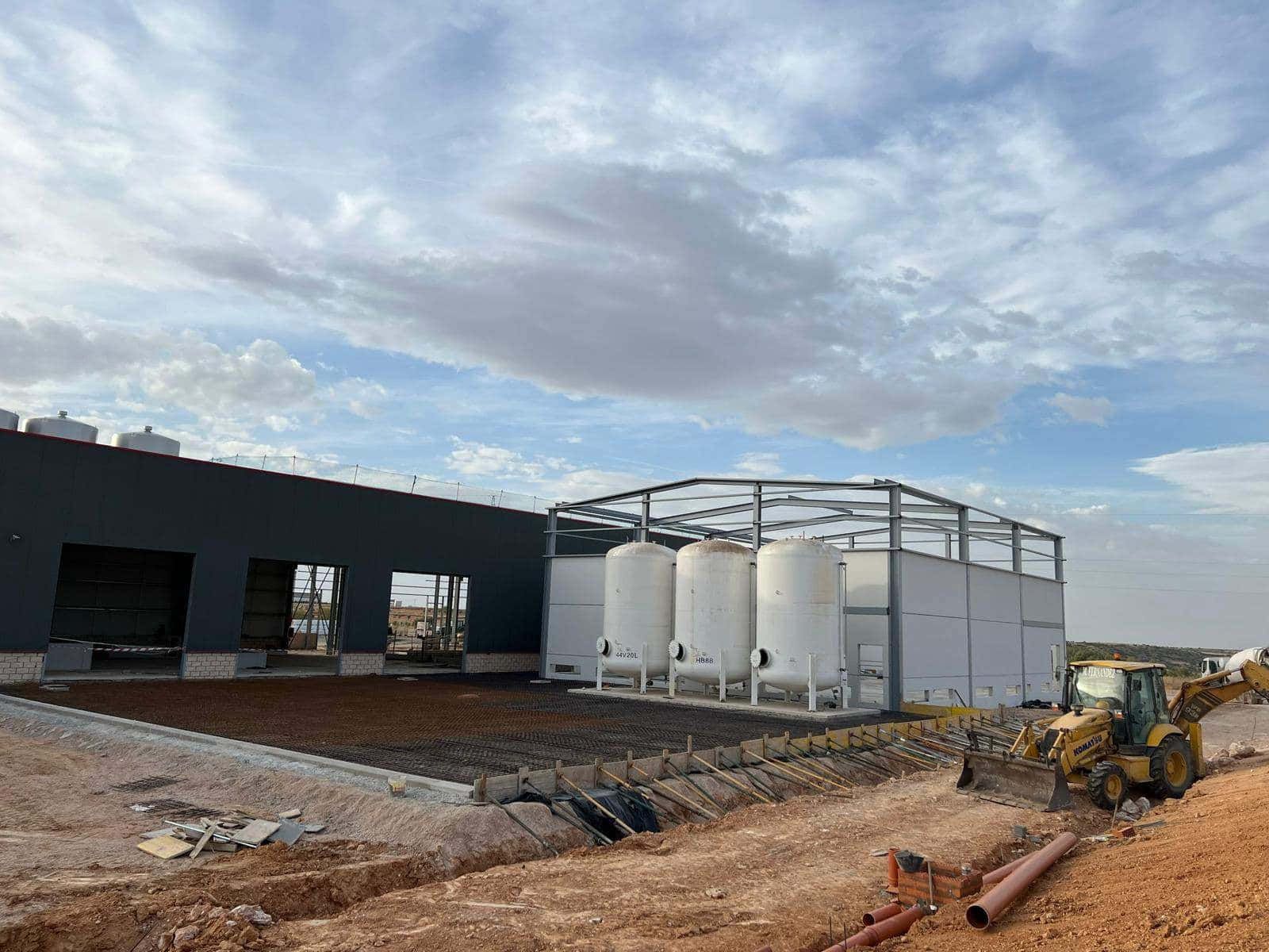 Sitio de construcción con un gran edificio gris, tanques blancos y un tractor amarillo bajo un cielo nublado.