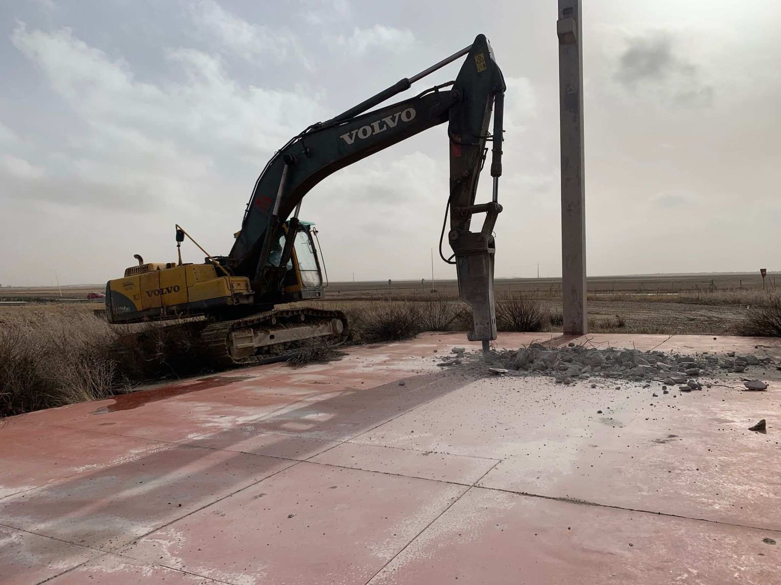 Una excavadora Volvo amarilla con un martillo neumático rompiendo hormigón cerca de un poste alto.