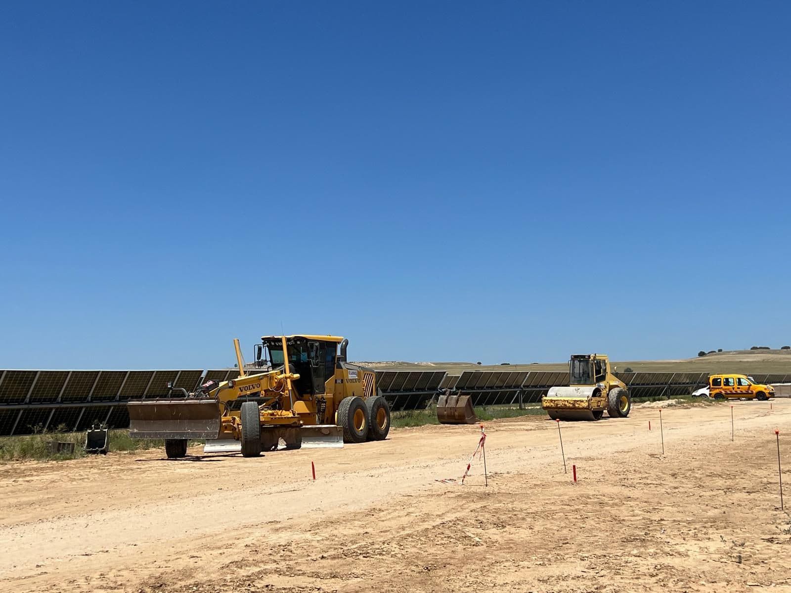 Vehículos de construcción nivelando un camino de tierra al lado de un conjunto de paneles solares bajo un cielo azul.