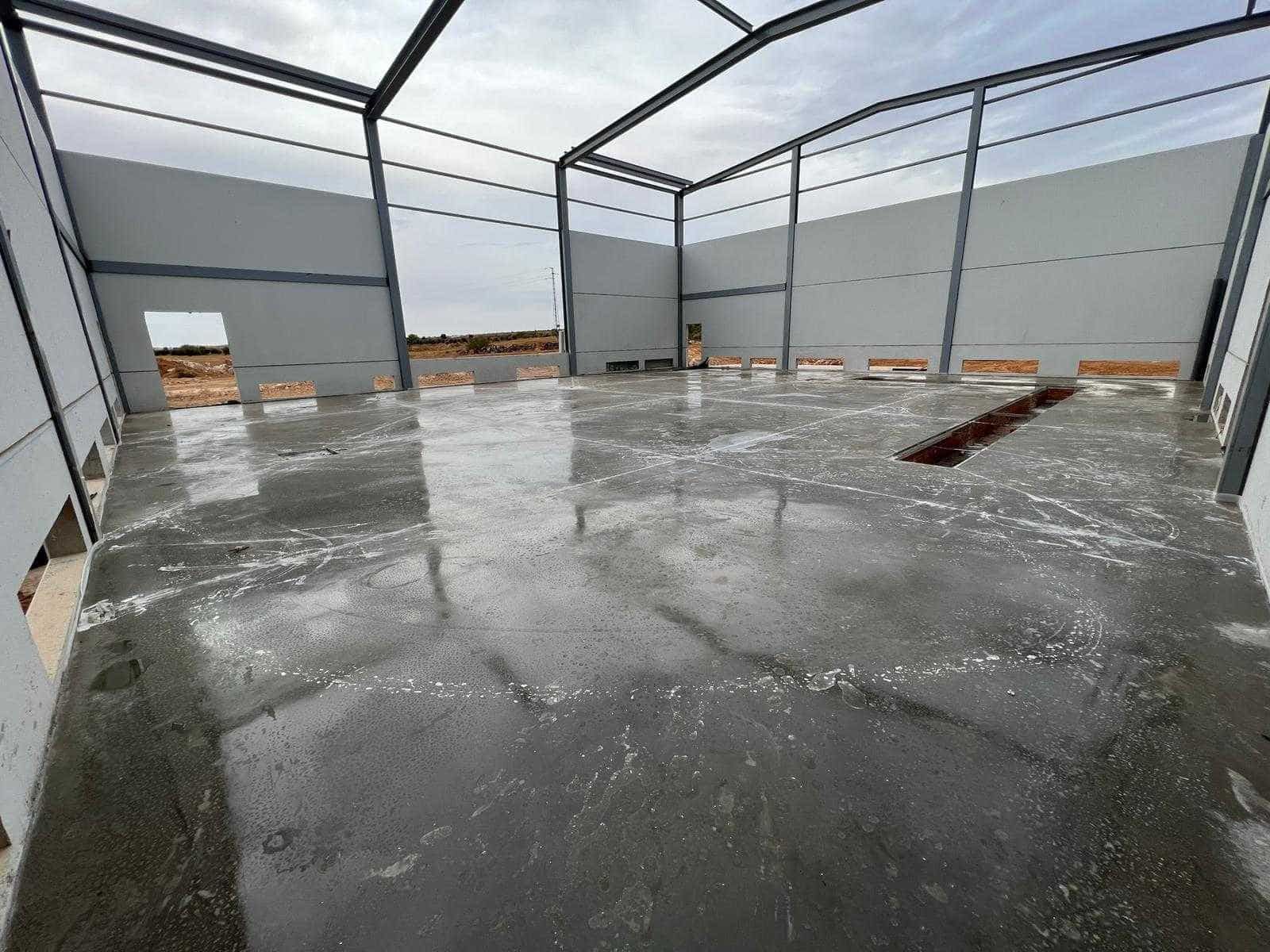 Interior de un gran edificio metálico con piso de concreto en construcción; paredes grises, estructura de acero.