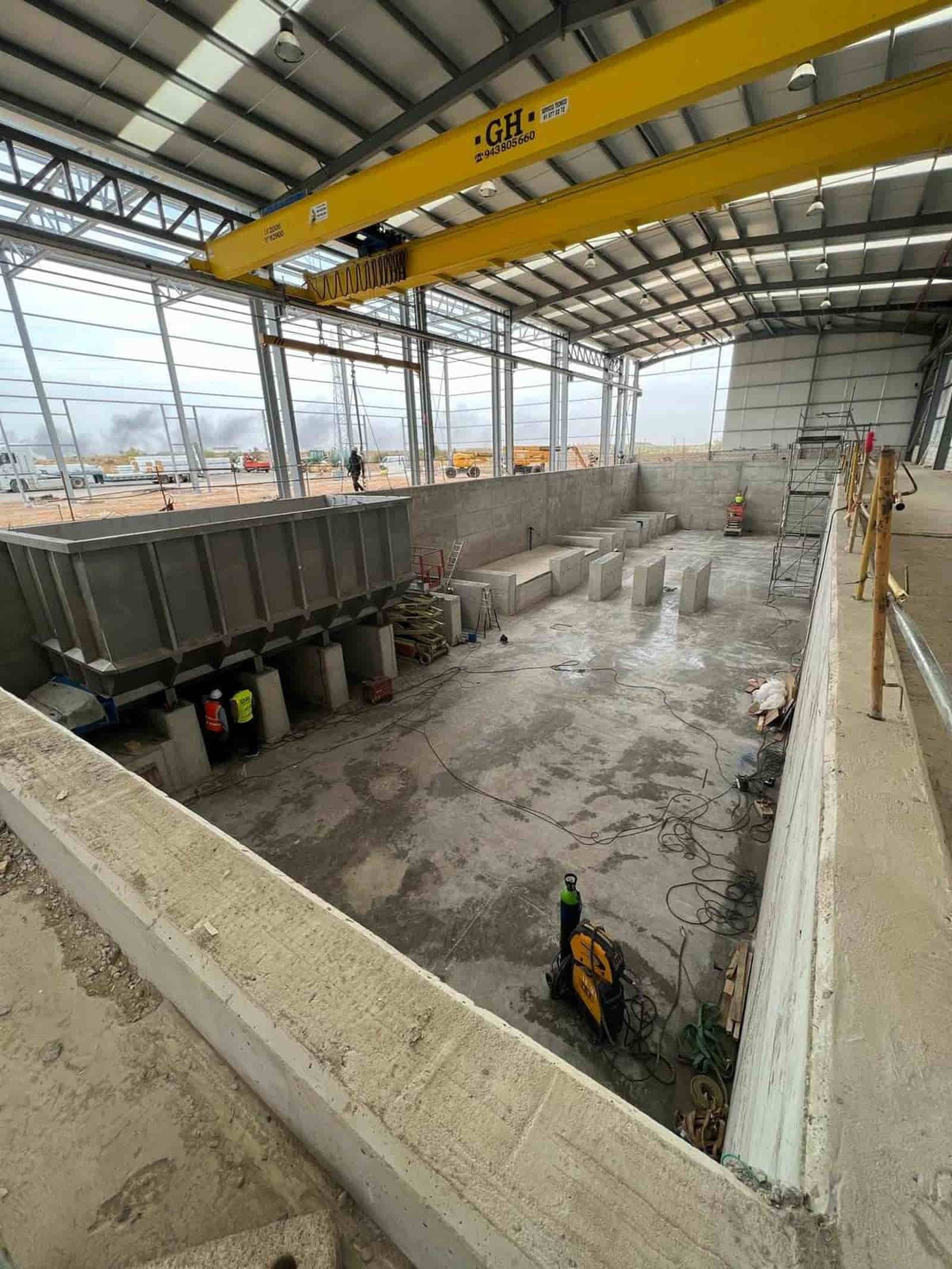 Interior de la obra; muros y pilares de hormigón. Grúa amarilla en lo alto. Trabajadores y maquinaria presentes.